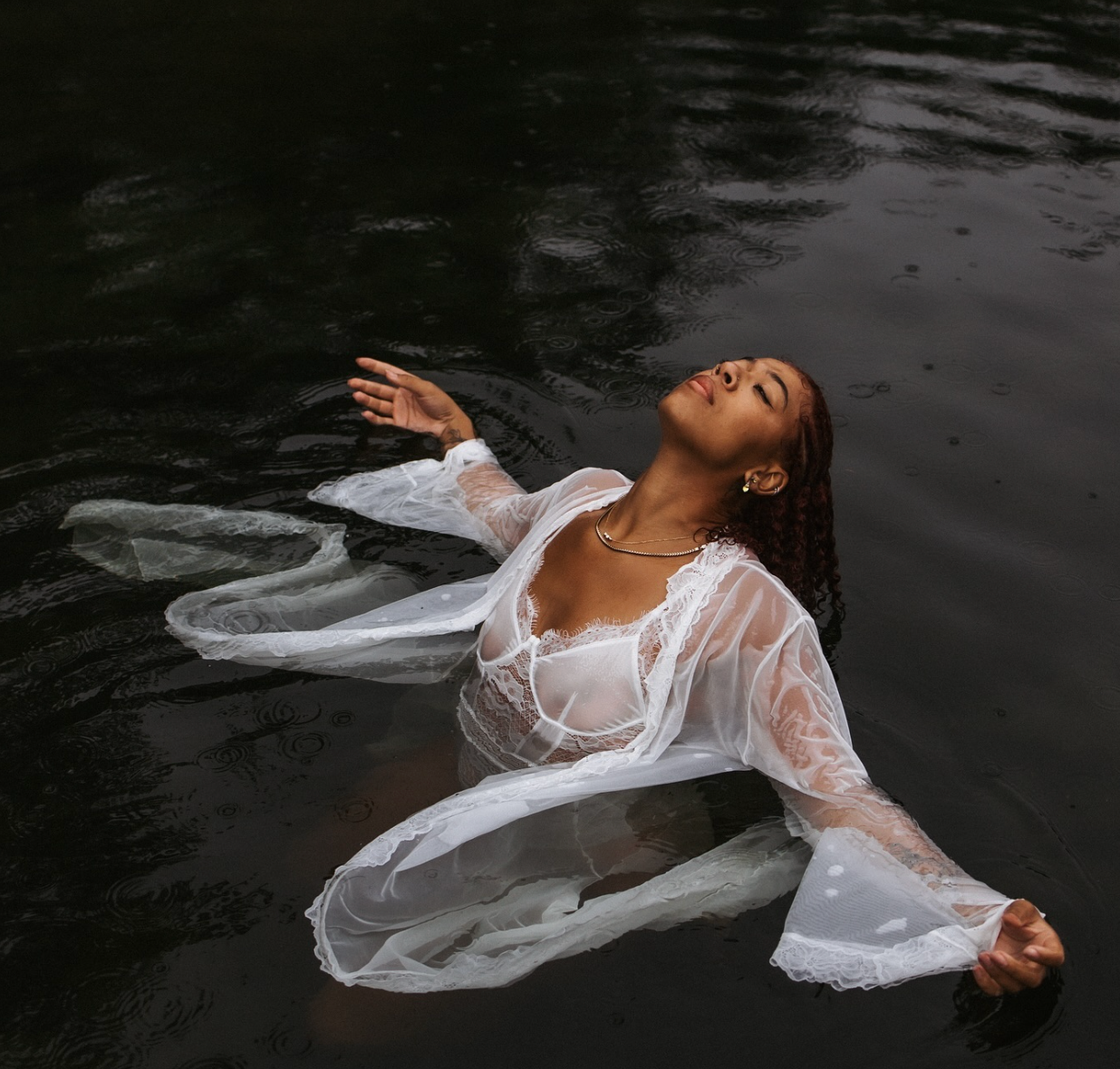 Artistic portrait of person floating in dark water wearing flowing white lace garment.
