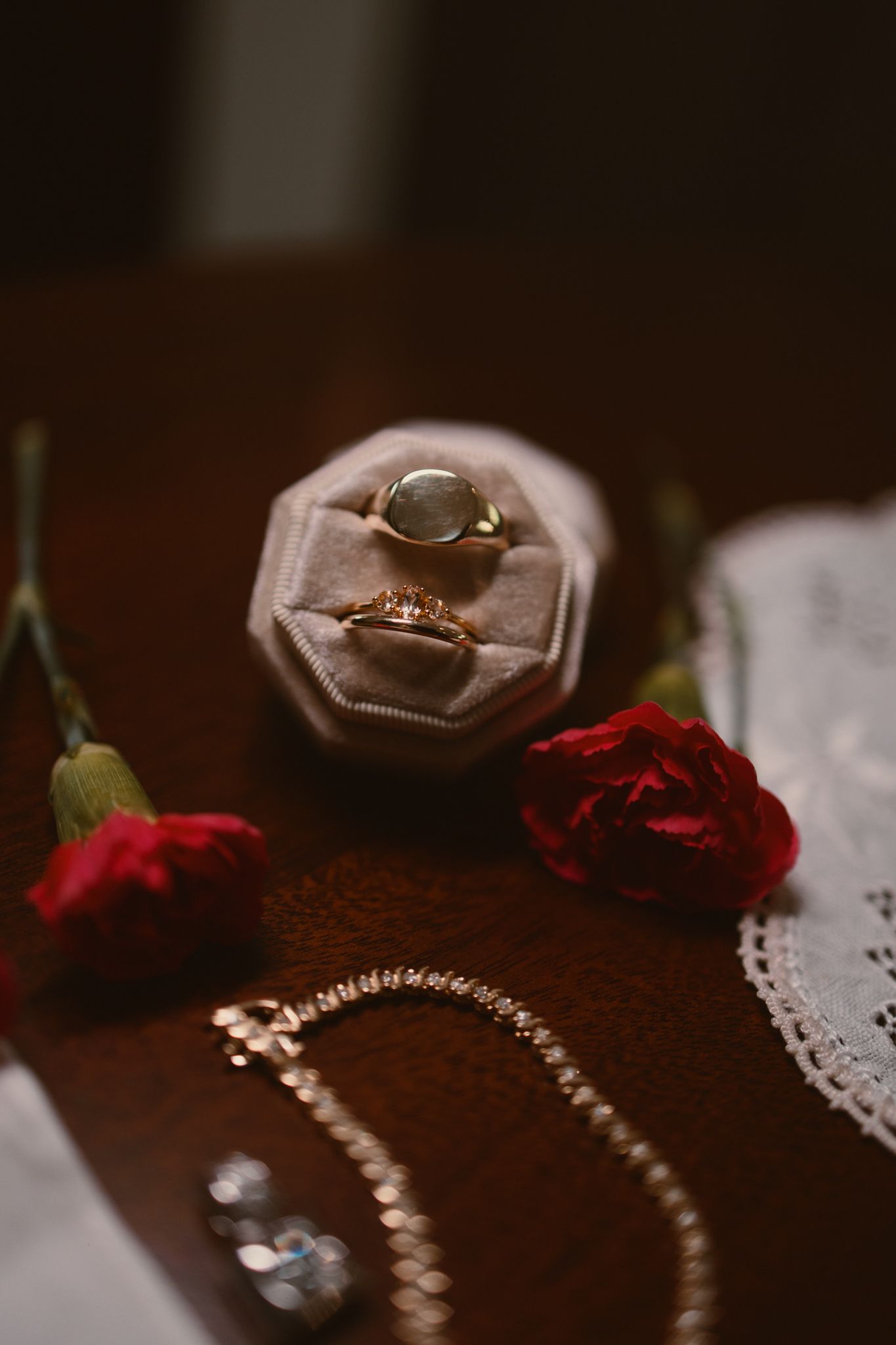 Wedding rings displayed on a plush ring holder with red roses and jewelry accents on a dark wooden surface.