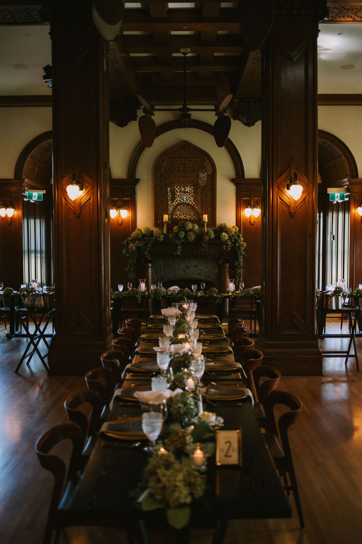 Elegant dining hall with dark wood pillars, warm lighting, and long banquet tables decorated with floral centerpieces.