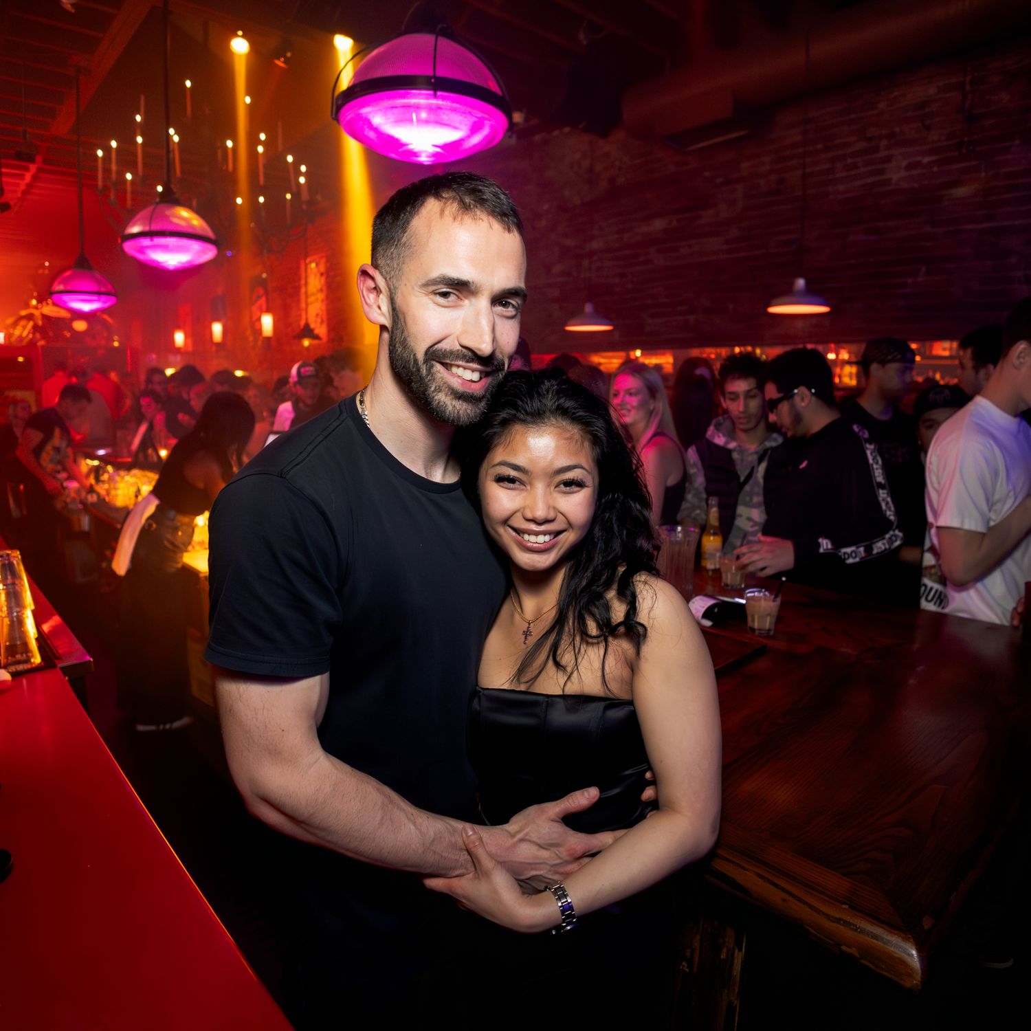 A couple embraces on a dance floor under pink lighting at a nightclub with other patrons visible in the background.