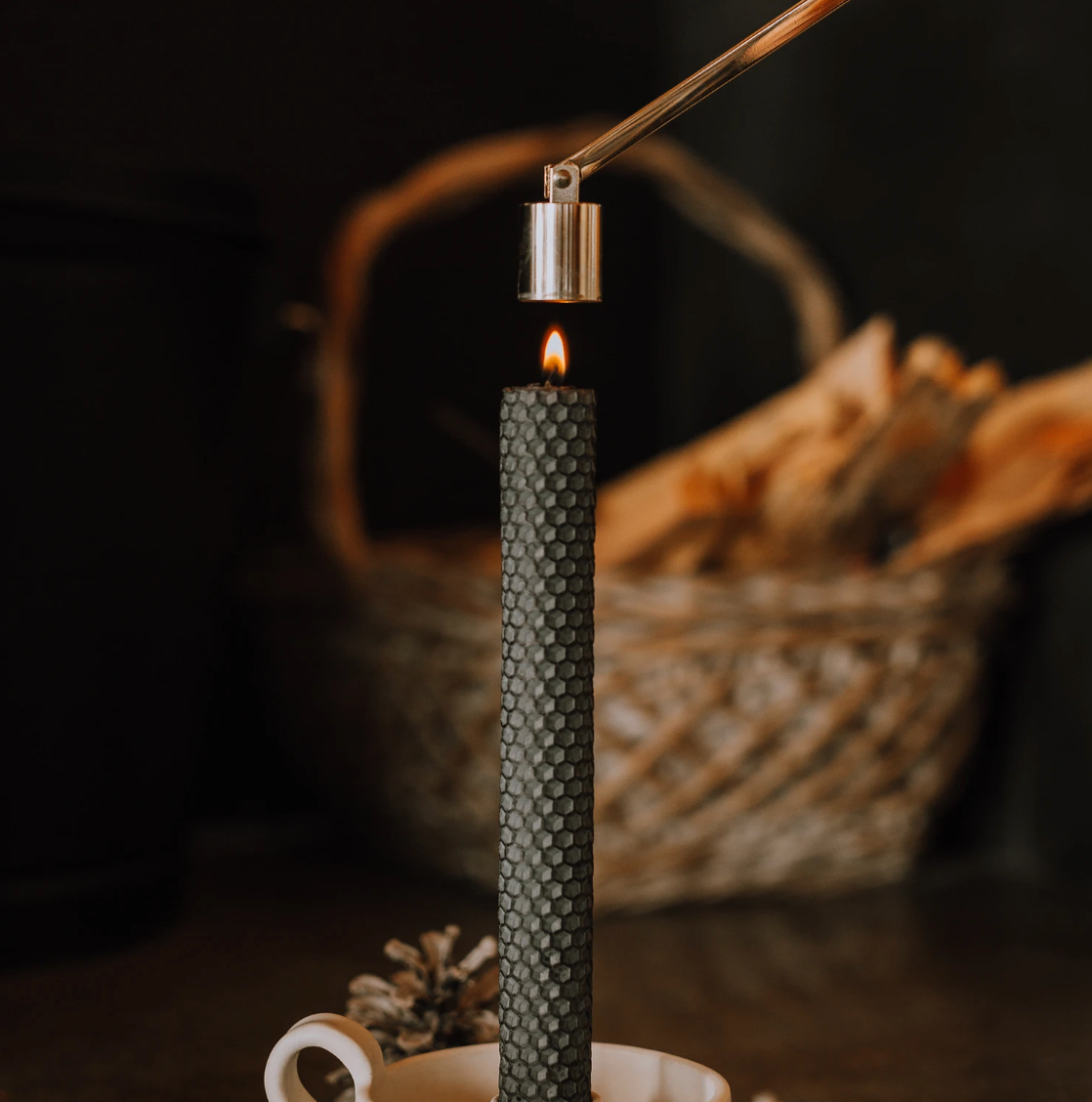 Black textured pillar candle burning on a white holder with pine cones in the background.