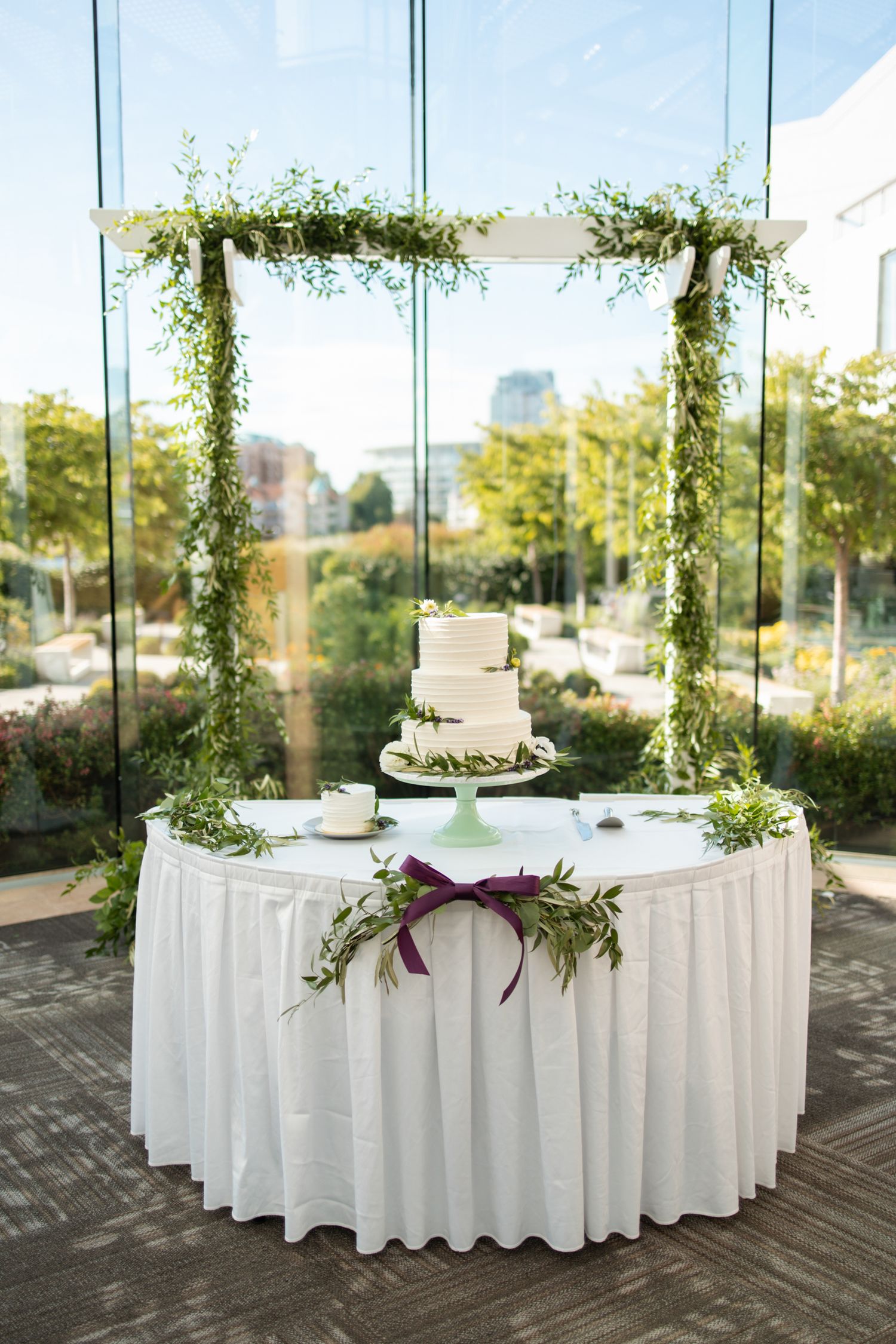 A minimalist wedding cake display on a white tablecloth with suspended greenery arch decoration.
