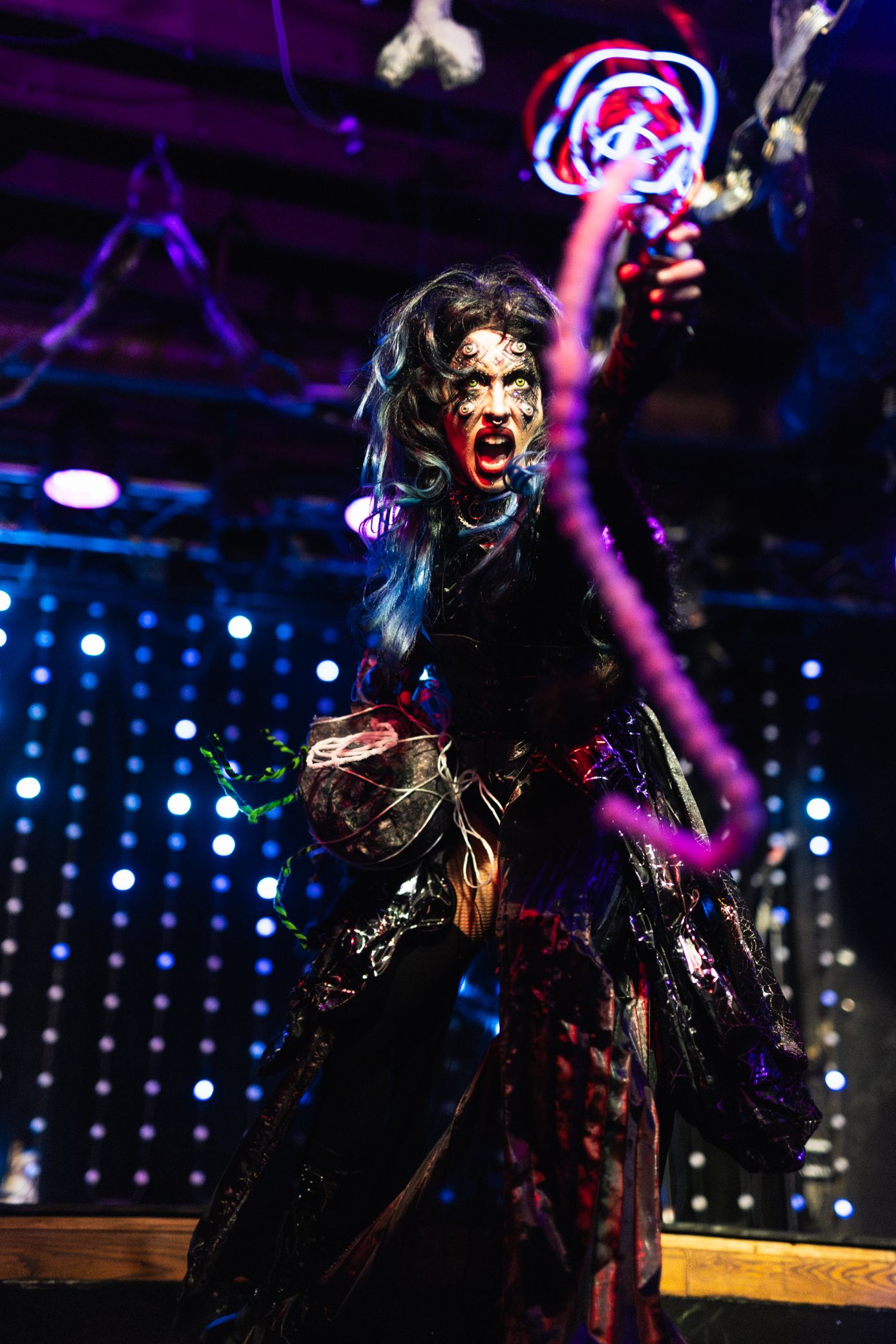 A performer in an elaborate dark costume and dramatic stage makeup poses energetically against a purple-lit stage backdrop.