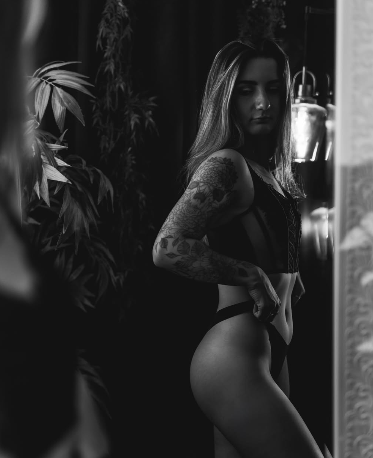 Black and white artistic portrait with tattoos and indoor plants in dramatic lighting.