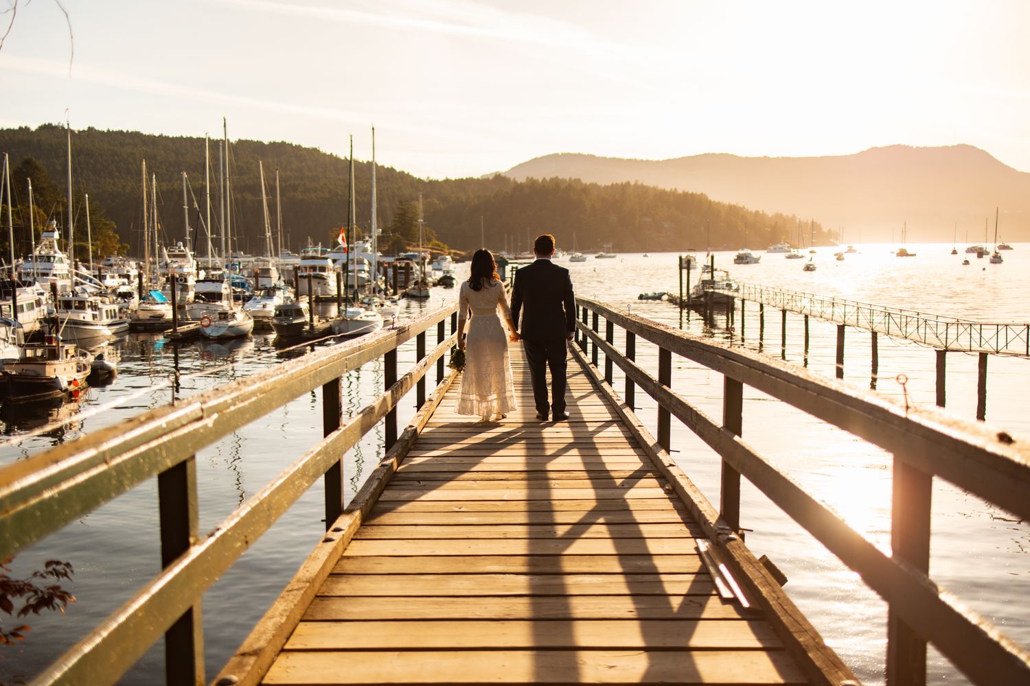 Silhouetted couple walking on wooden dock at sunset with boats in marina background.