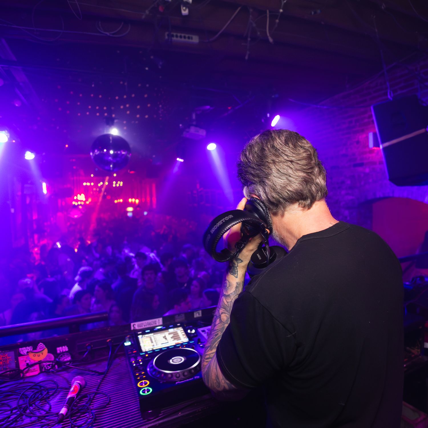 DJ performing at a nightclub with vibrant purple lighting and crowd in background during a live music event.