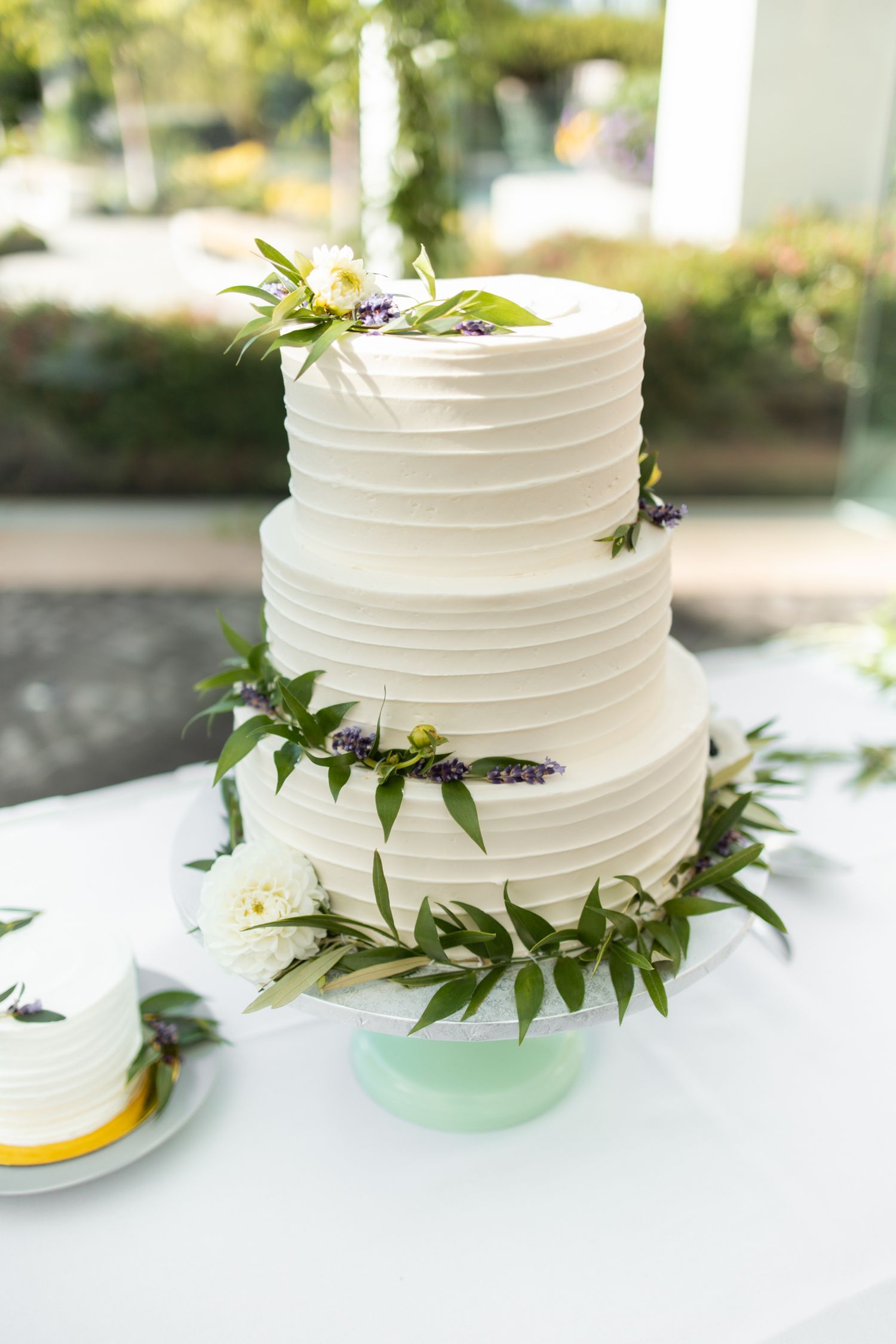Three-tiered white wedding cake decorated with delicate horizontal textures and fresh greenery on a mint green cake stand.