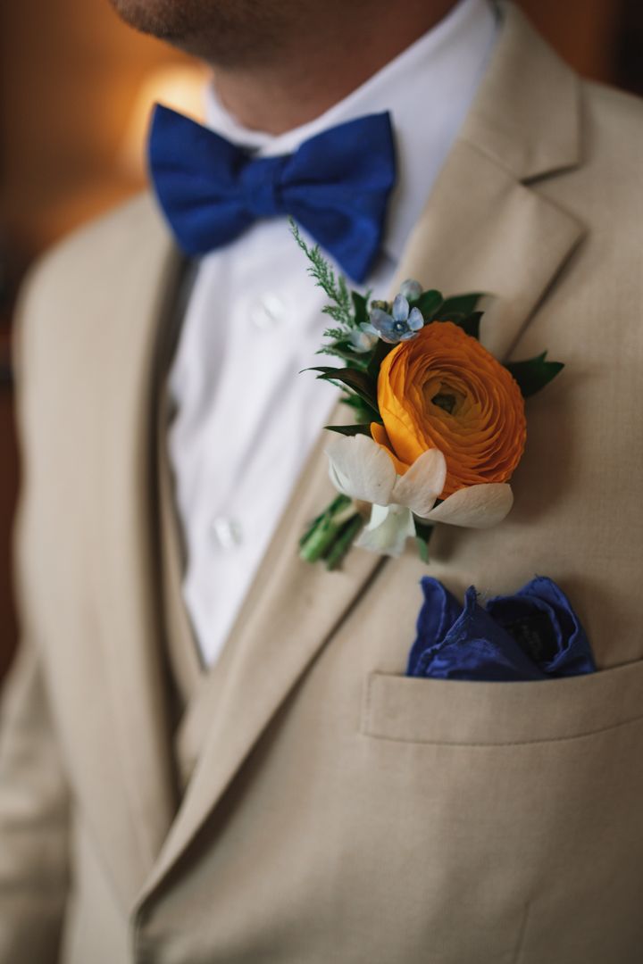A beige suit jacket with navy blue bow tie and pocket square features an orange ranunculus and white flower boutonniere.