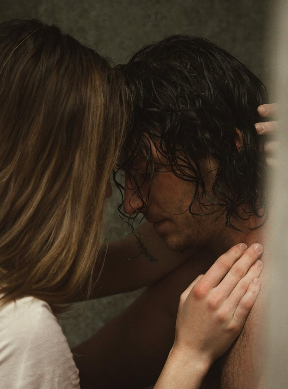 Intimate portrait of couple sharing tender moment against textured wall in dim lighting.