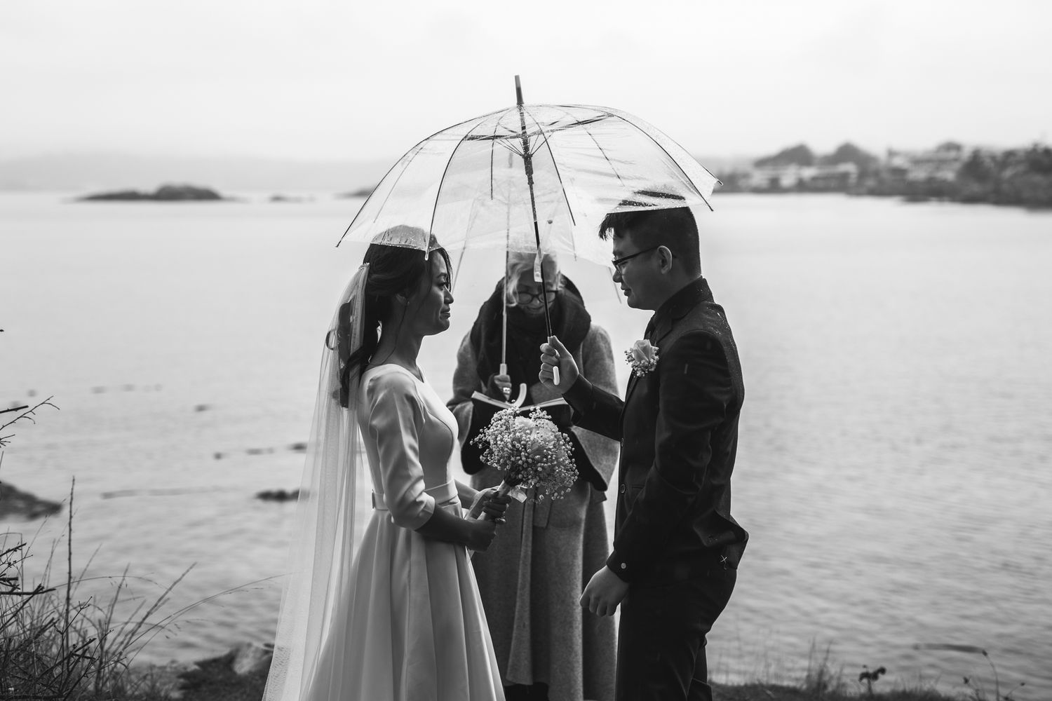Black and white wedding photo with couple holding umbrella together by the ocean during coastal ceremony.