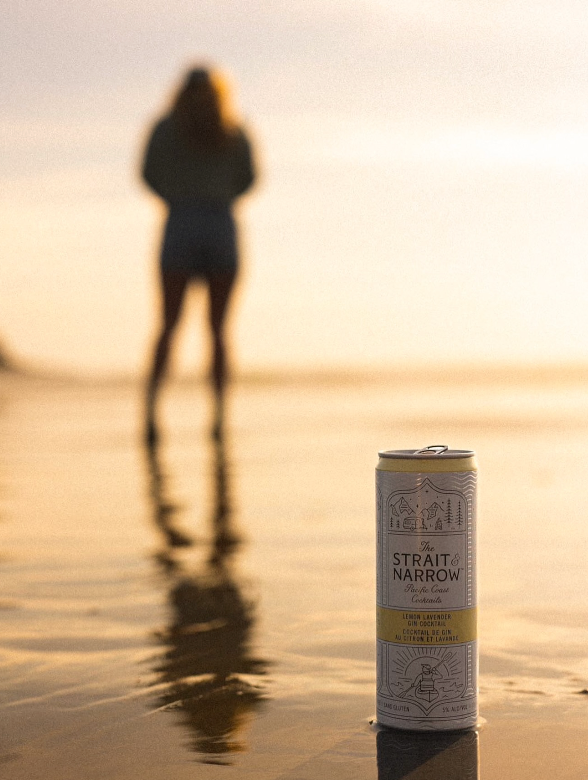 A silhouetted figure on a beach at sunset with a Strait and Narrow beverage can in the foreground.