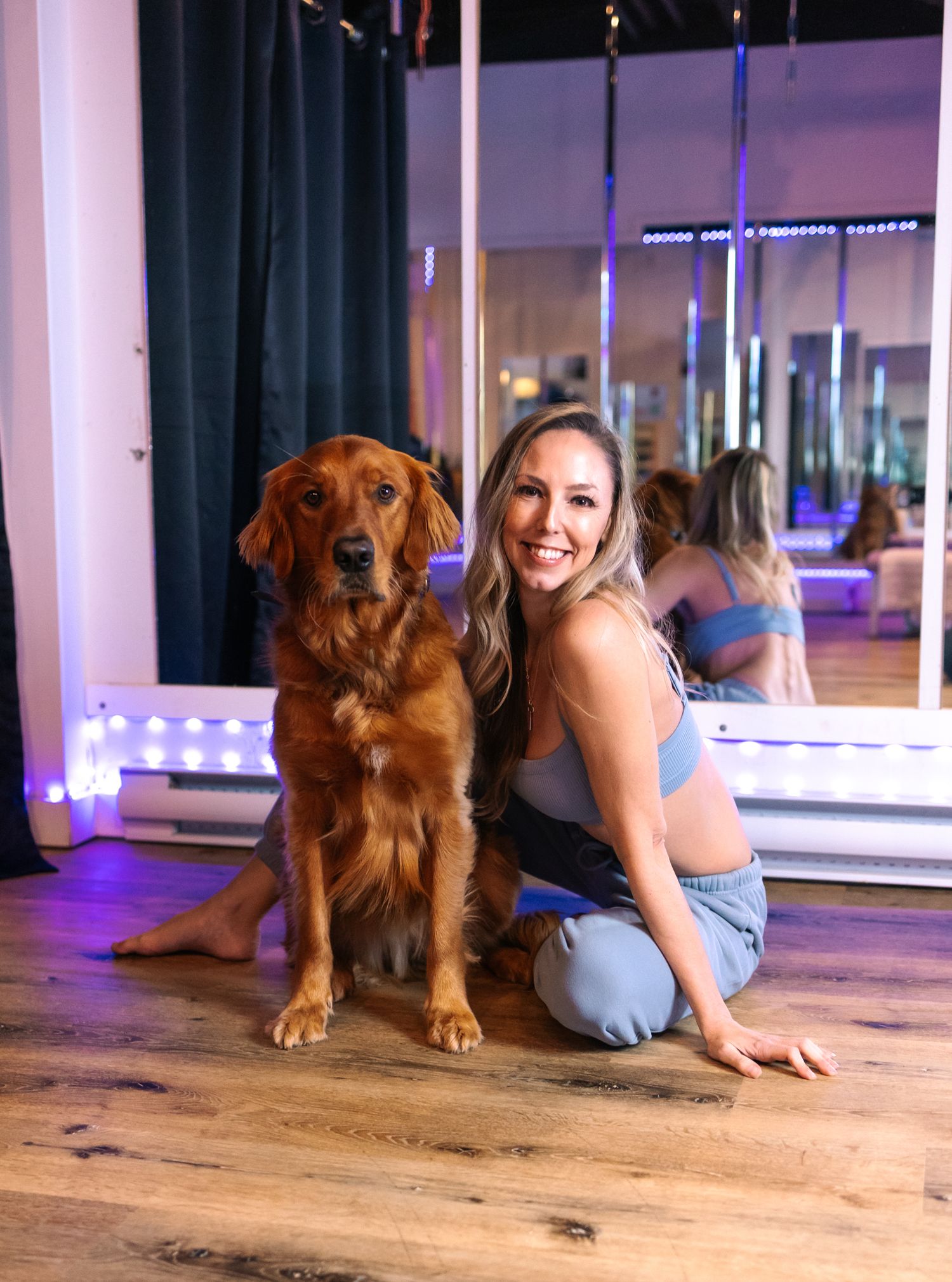 Golden retriever dog sits with woman in athletic wear on wooden floor of fitness studio with purple lighting.