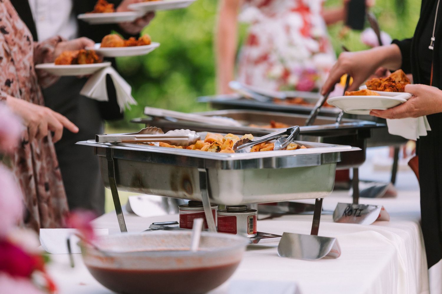 Buffet style catering setup with stainless steel chafing dishes and serving utensils at an outdoor event.