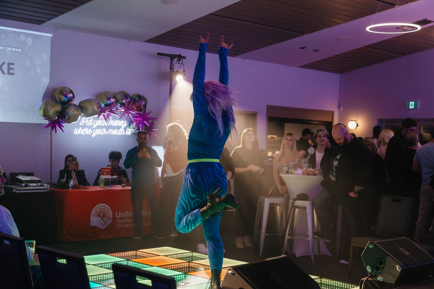 A breakdancer performs an impressive handstand on a lit-up platform in a dimly lit venue with purple lighting.