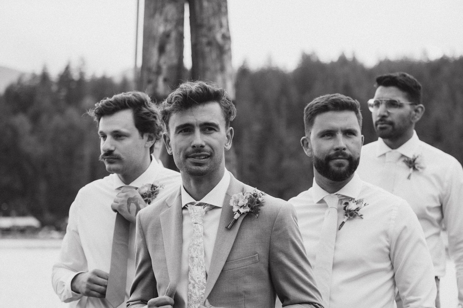 A black and white photo shows groomsmen wearing suits and boutonnières standing together outdoors.