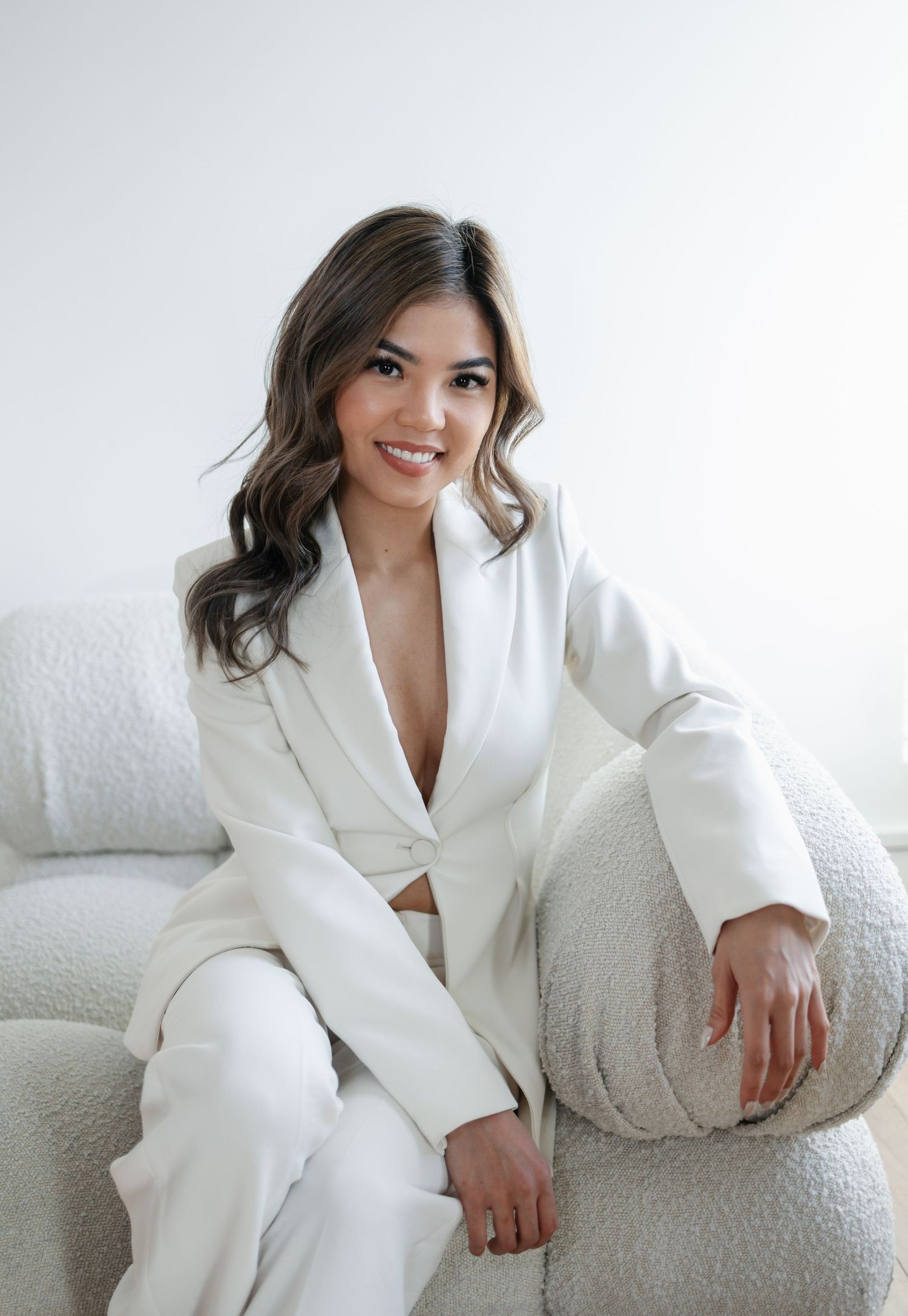 Professional fashion photo of model in white tailored suit sitting on white couch.