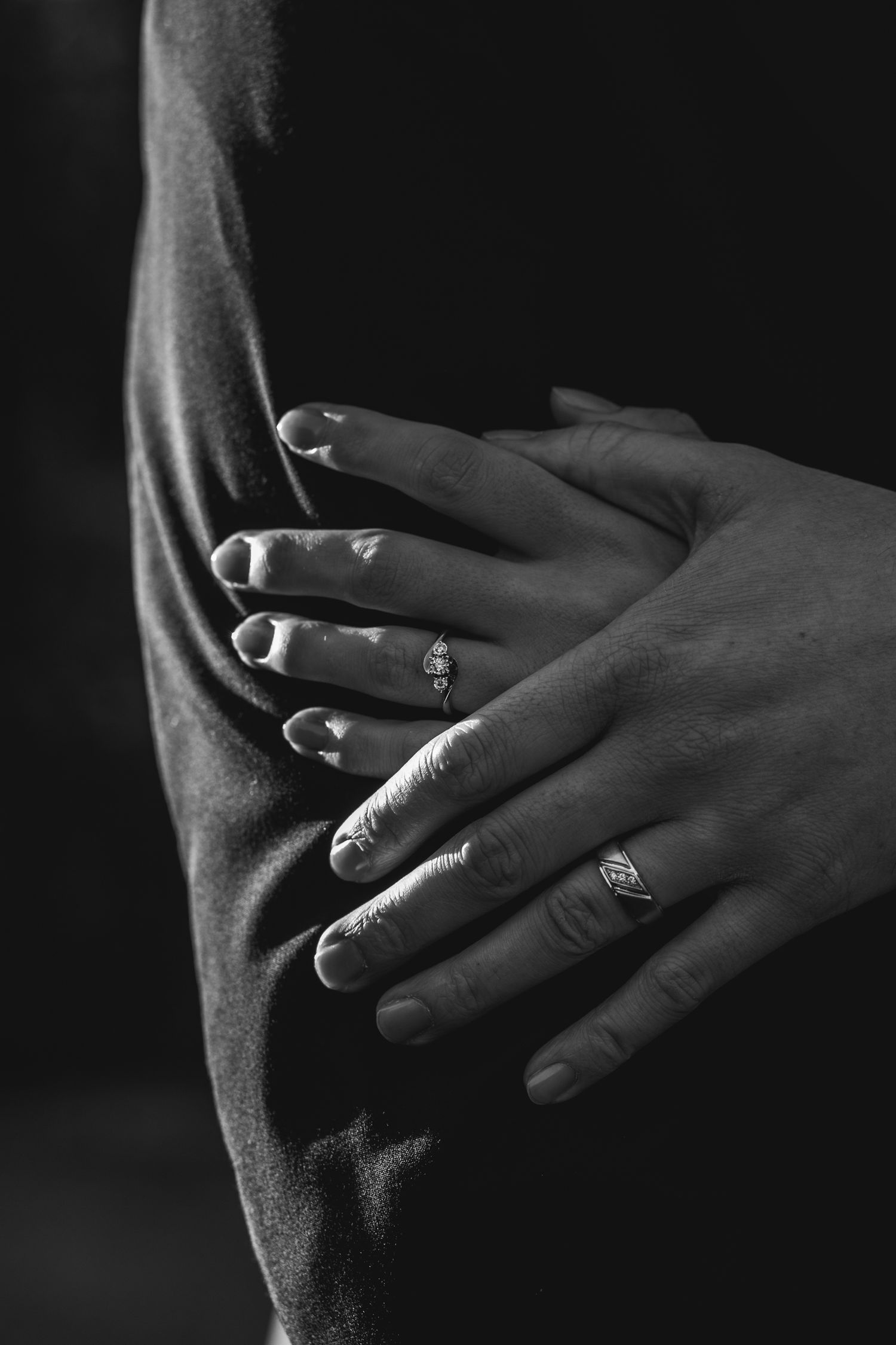 Black and white close-up of hands with wedding rings against dark fabric.