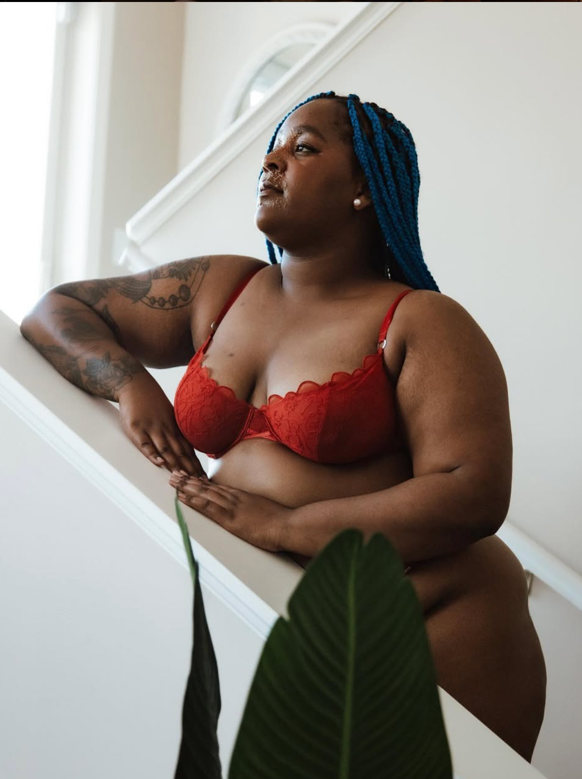 Body positive portrait featuring red lingerie and blue braids against white wall with tropical plant.