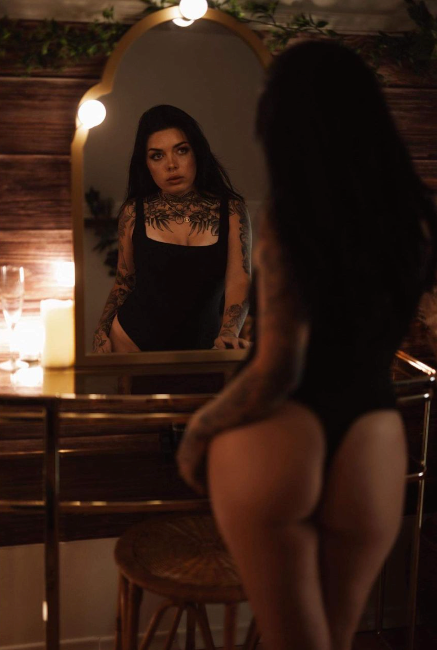 Person in black bodysuit looking at reflection in candlelit mirror with wooden frame.