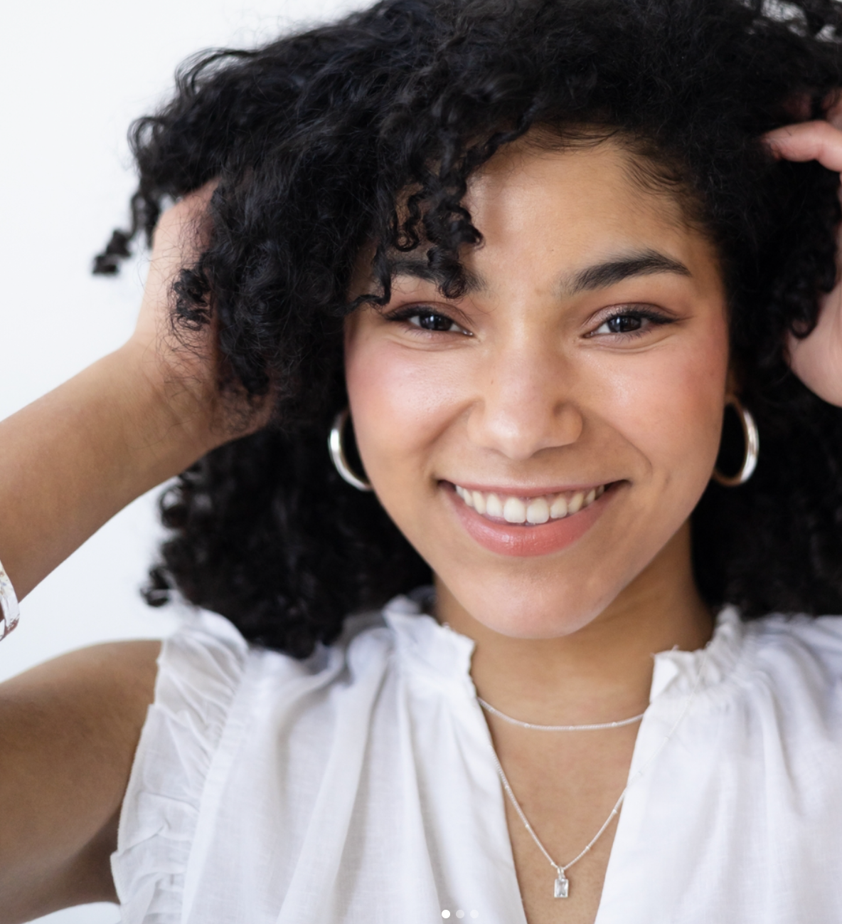 A close-up portrait showing natural curly hair, a bright smile, and delicate layered necklaces against a light background.