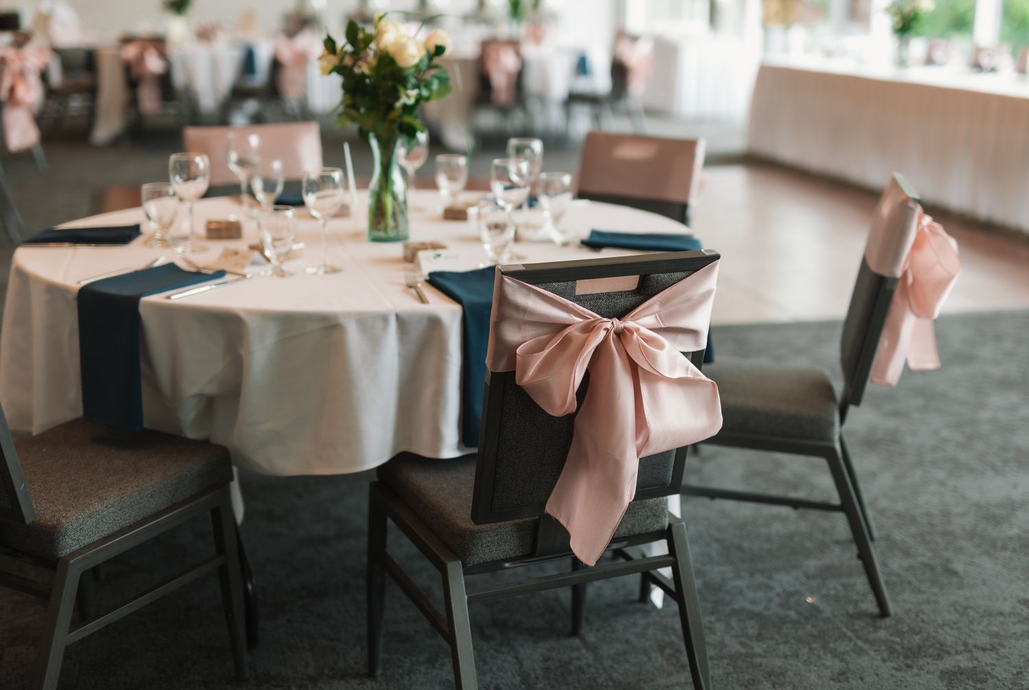 A wedding reception table setup with white linens, pink bow chair decor, and fresh floral centerpieces.