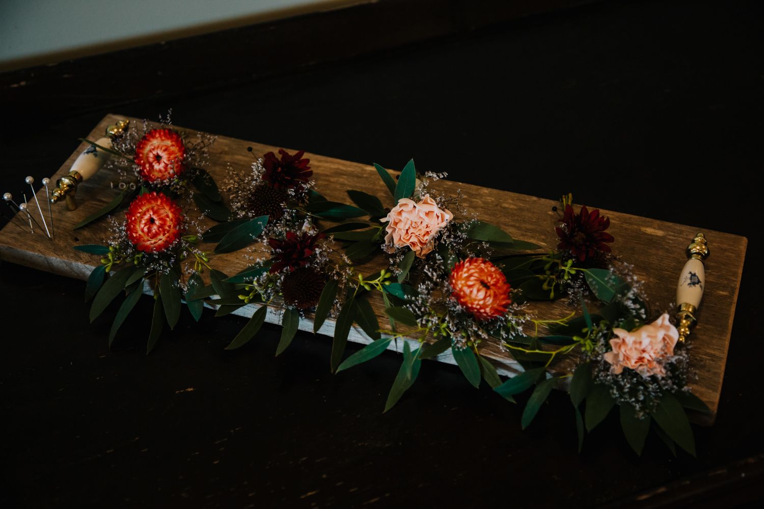 A rustic wooden board adorned with a vibrant floral arrangement featuring orange dahlias and greenery against a dark background.