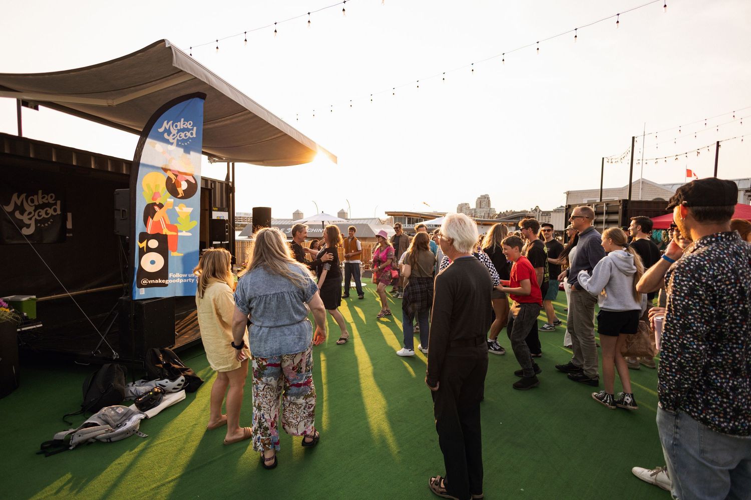 People gather at an outdoor event venue with astroturf flooring and string lights at sunset.