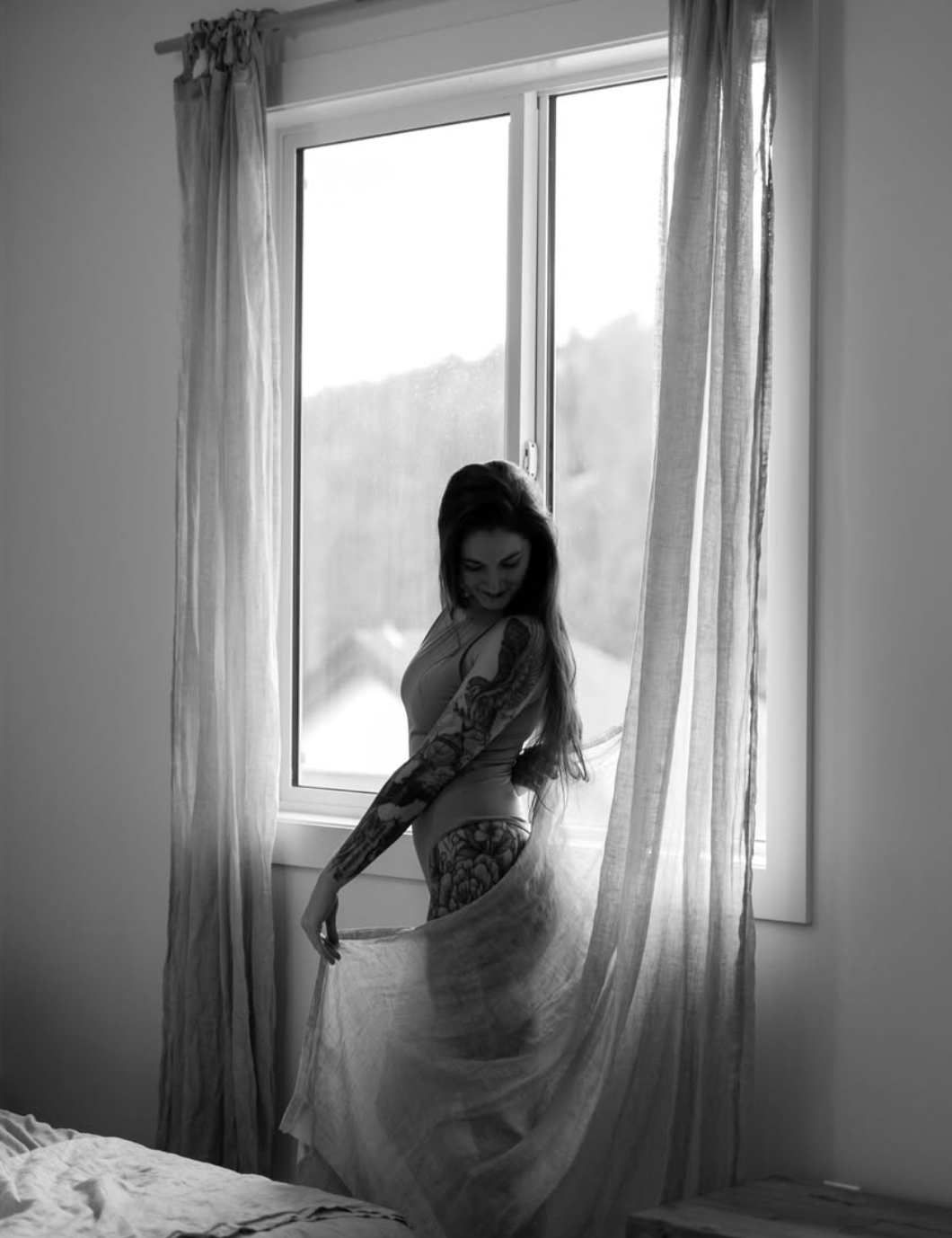 Artistic black and white boudoir photo by a window with flowing curtains.