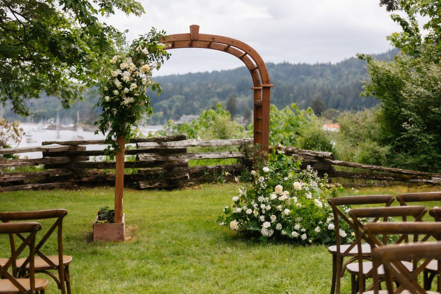 A rustic wooden wedding arch decorated with white flowers overlooks a mountain view with wooden guest chairs.