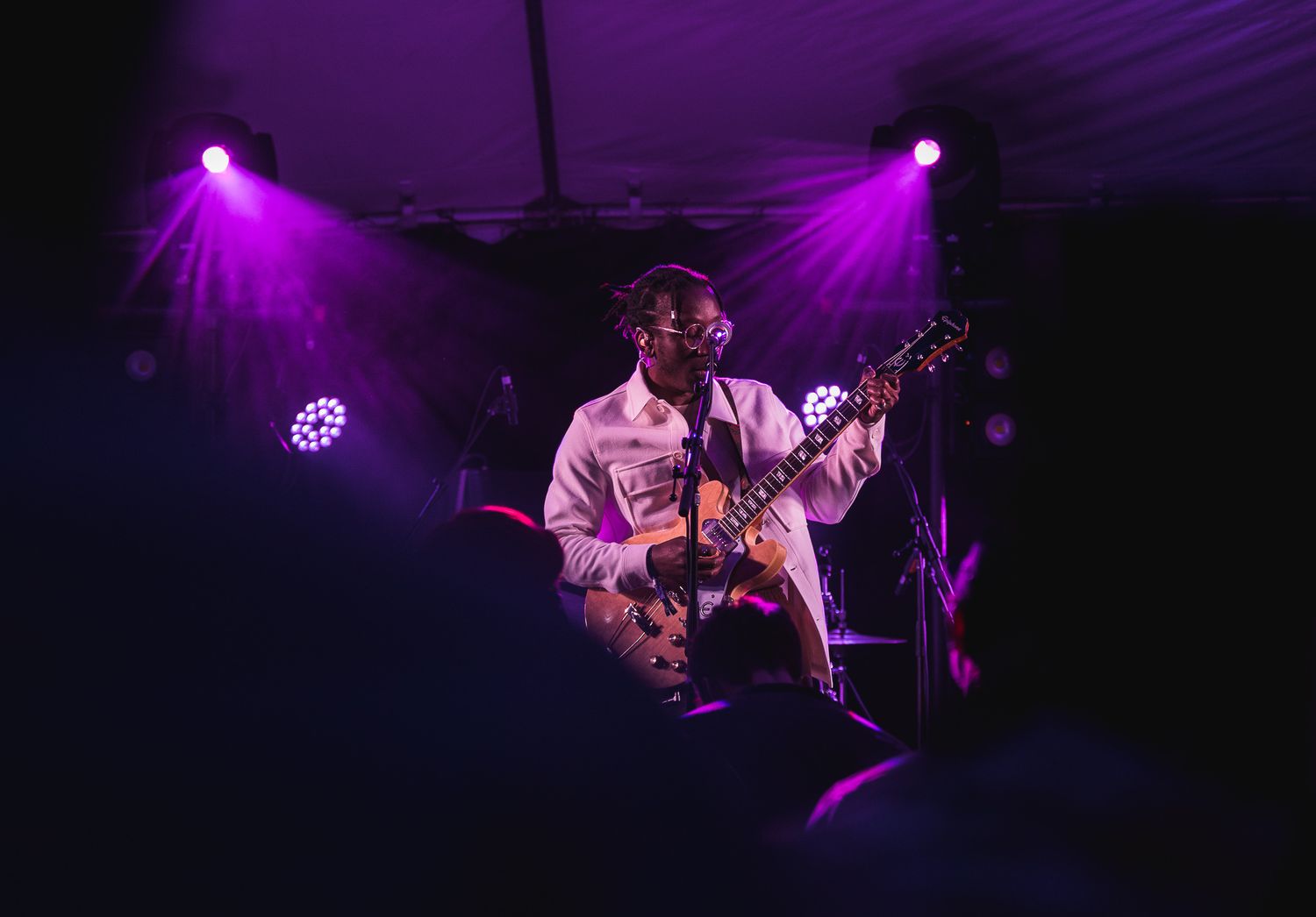 Performer in white shirt plays guitar on dimly lit stage with dramatic purple spotlights creating atmospheric ambiance.