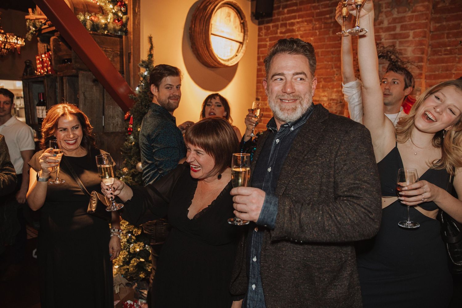 A group of guests enjoy drinks and laughter at an elegant indoor celebration with exposed brick walls and mood lighting.
