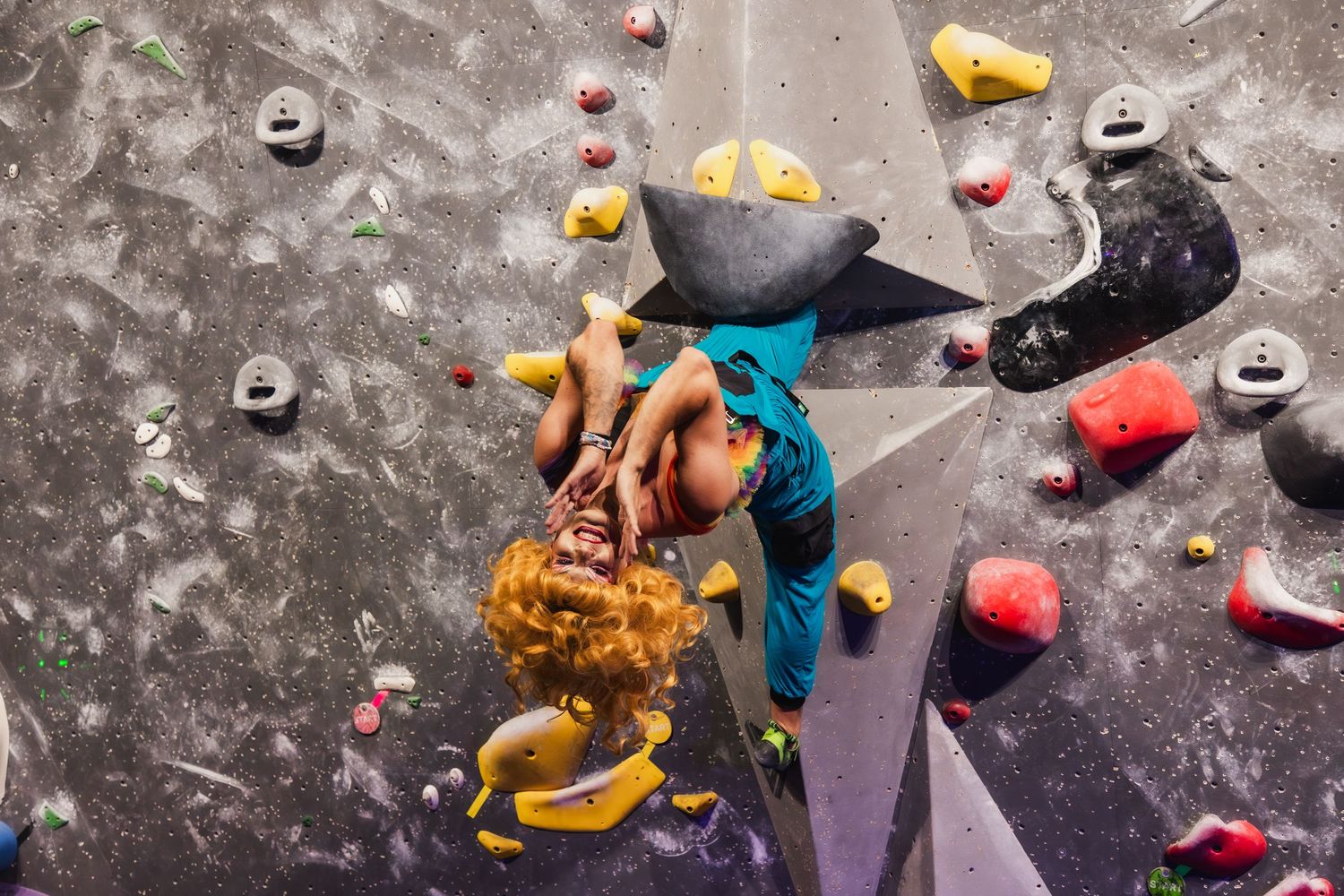 Rock climber in blue shirt scaling an indoor climbing wall with colorful holds and chalk marks visible.