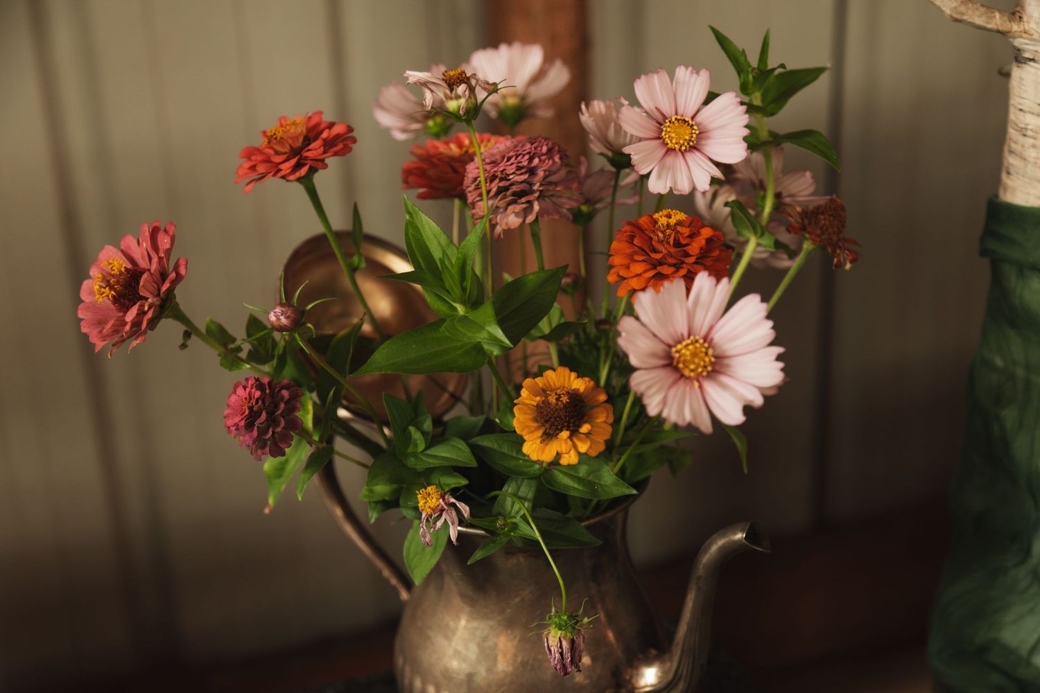 Vintage teapot arrangement with coral and pink zinnias and wildflowers in moody lighting.
