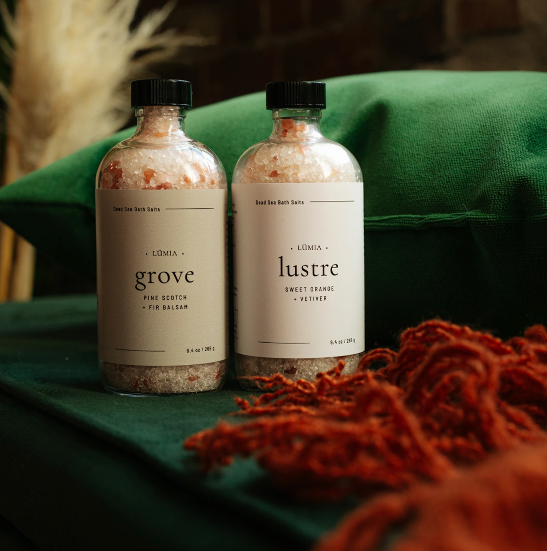 Two minimalist skincare bottles labeled 'grove' and 'lustre' on green fabric with red tassels.