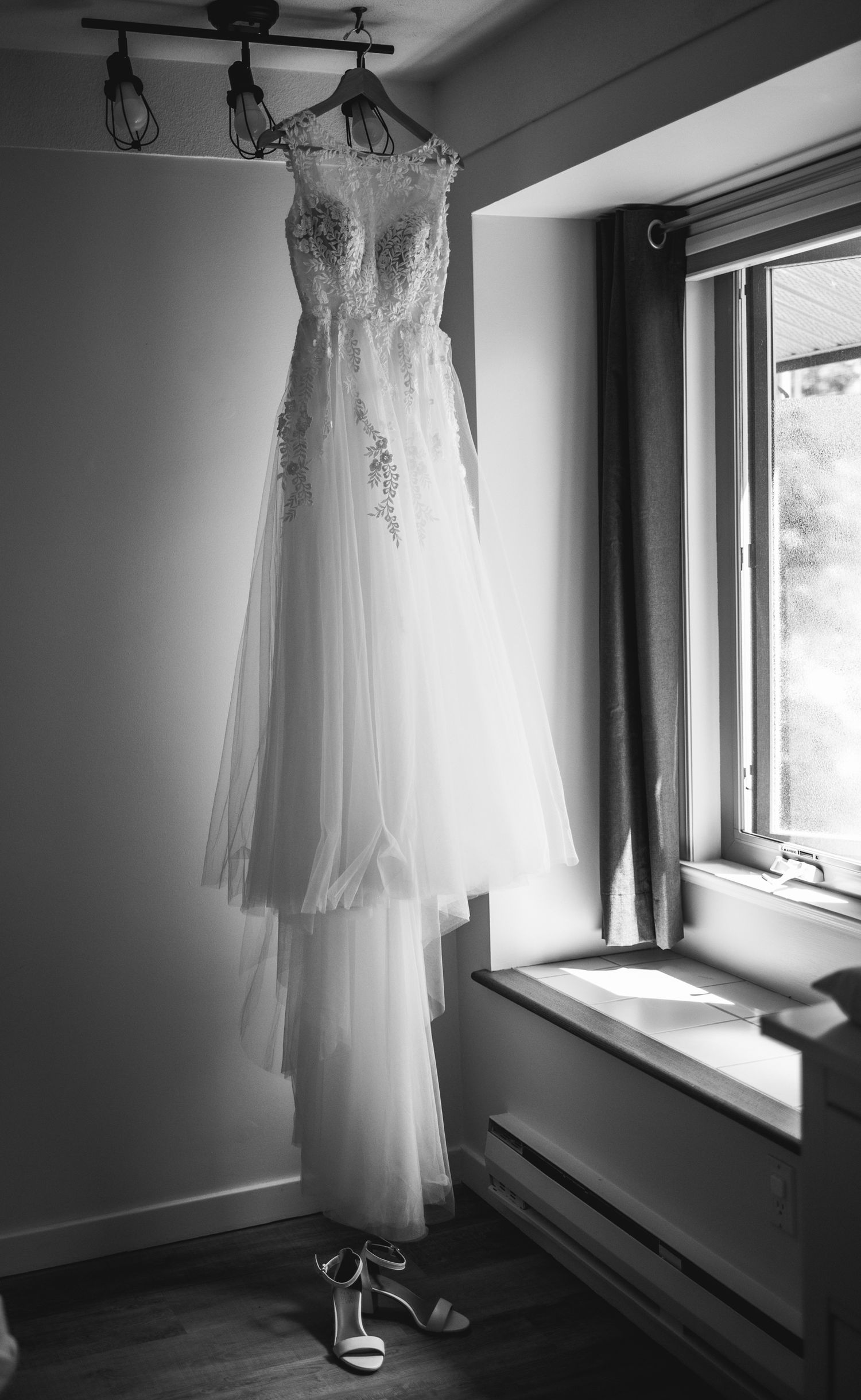 A delicate white wedding dress with lace details and embellishments hangs by a window, with matching shoes beneath.
