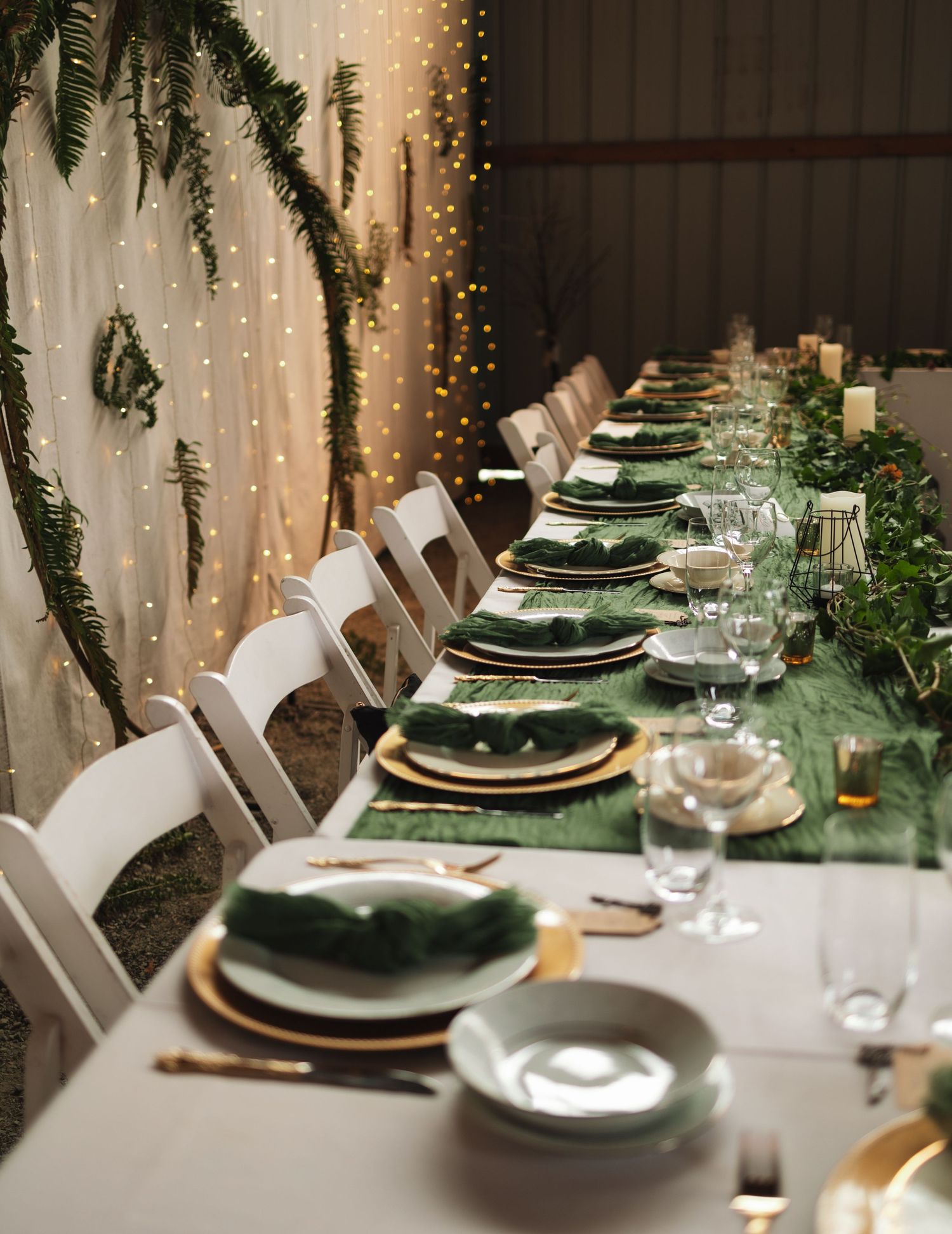 An elegant table setting with green and gold accents, white chairs, and twinkling fairy lights.