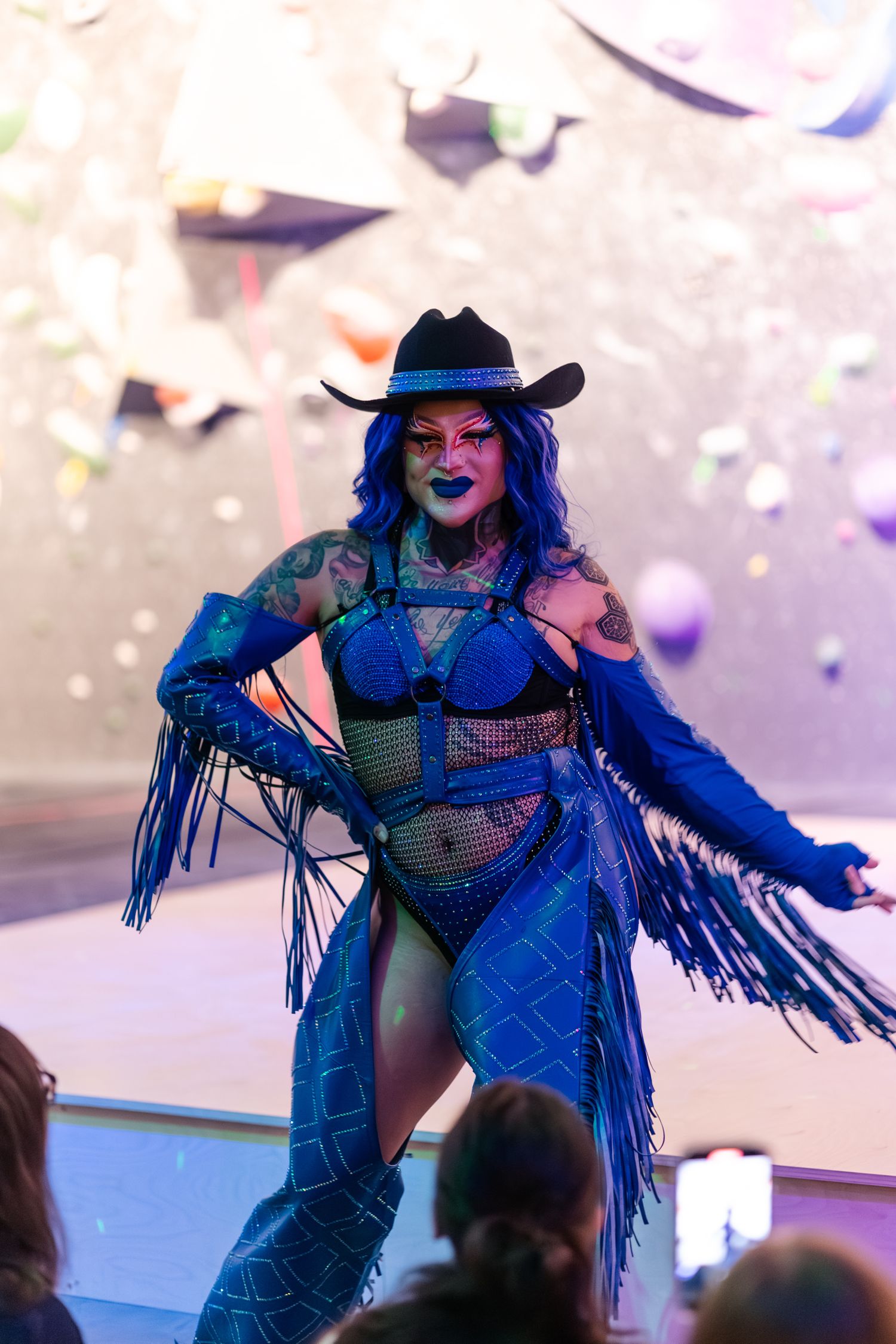 A performer in a blue fringe outfit and black cowboy hat dances on stage during a burlesque show.