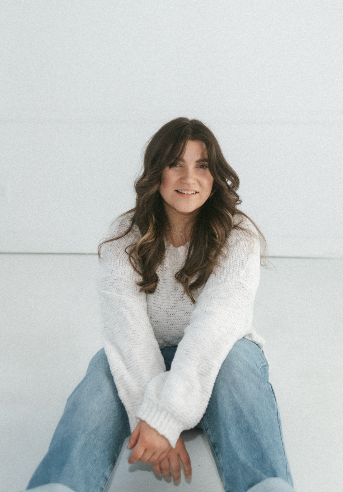 Person wearing a white knit sweater and blue jeans sits on floor against white wall in casual pose.