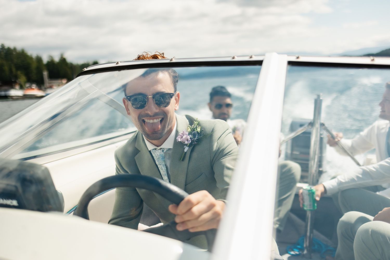 A person in sunglasses smiling while driving a boat on a sunny day near the shoreline.