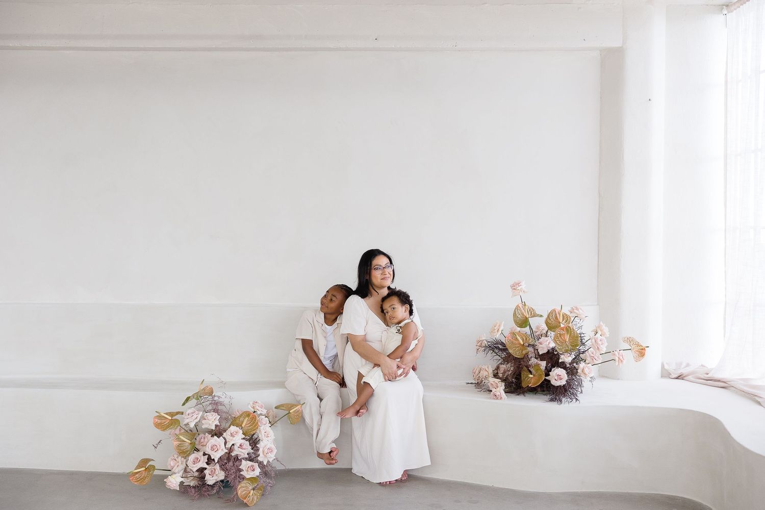 Minimalist white studio with scattered pink and white floral arrangements on a concrete floor beside person in flowing dress.