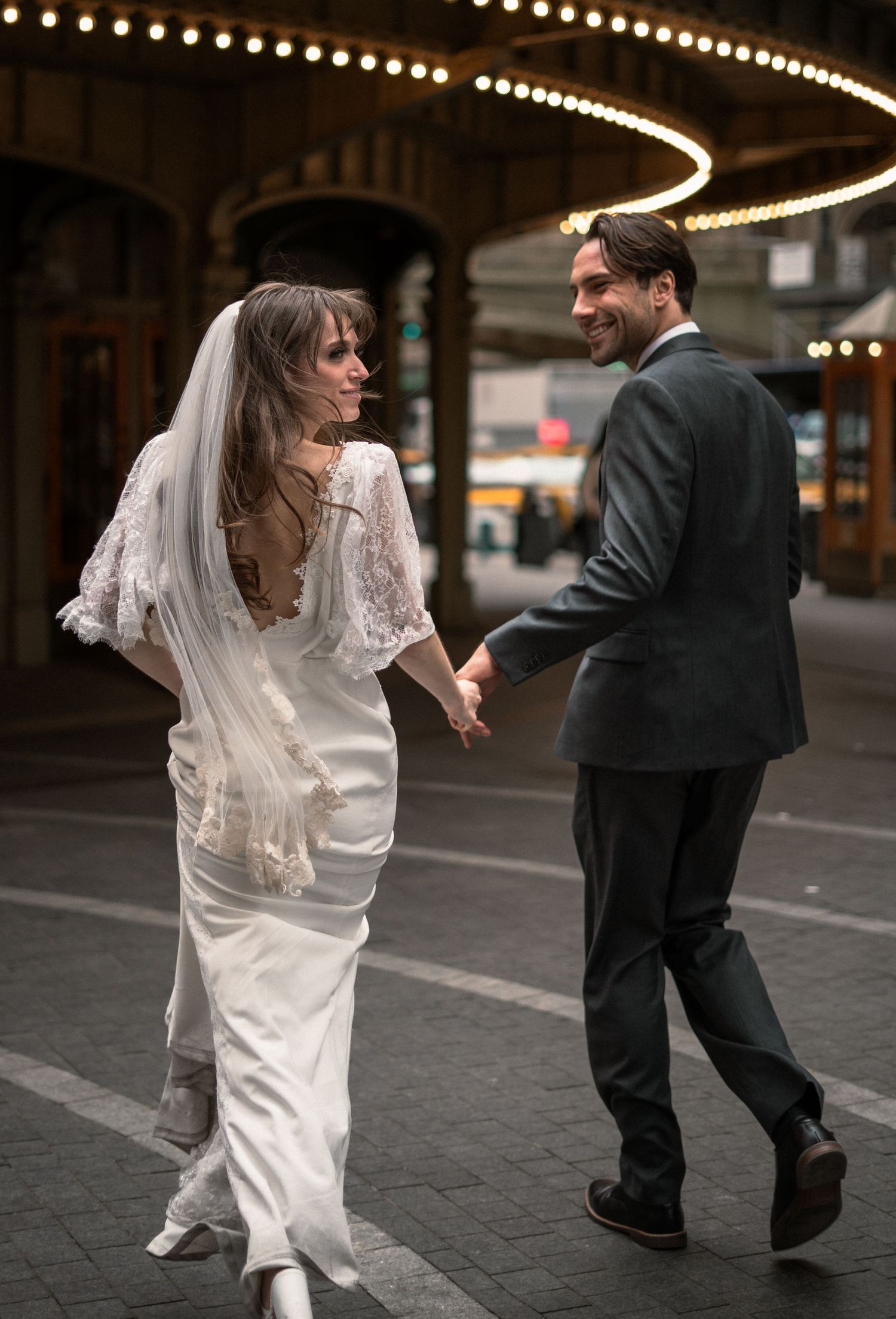 Newlywed couple walking and laughing together under string lights on city street at dusk in wedding attire.