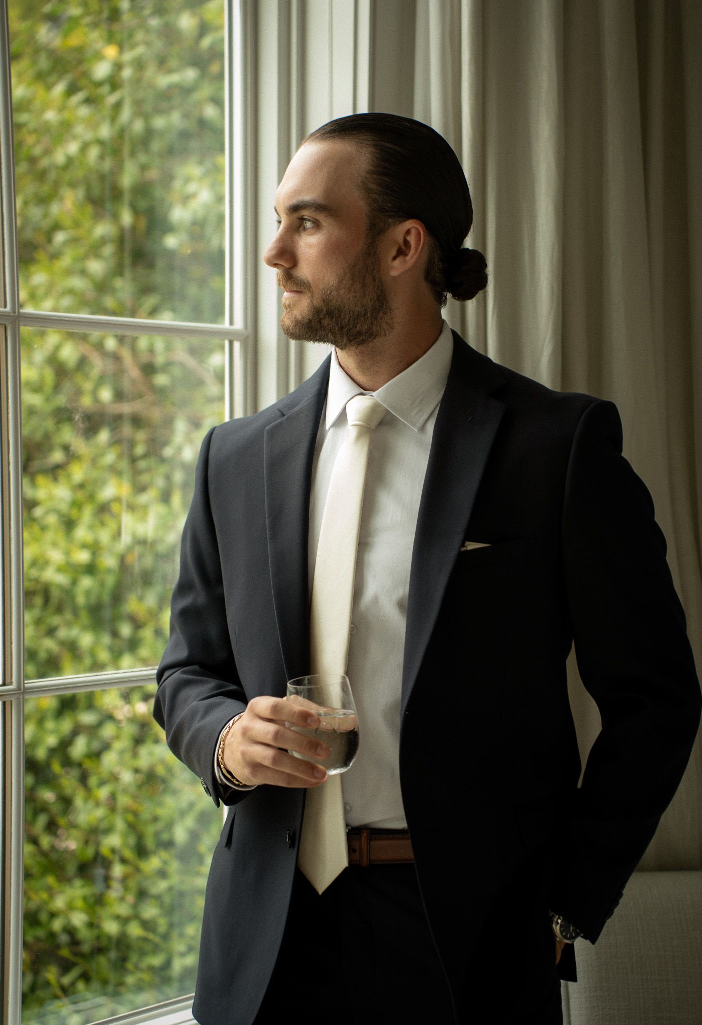 Groom standing by a window, sipping a drink and gazing out at the vineyard grounds in Hamptons, NY