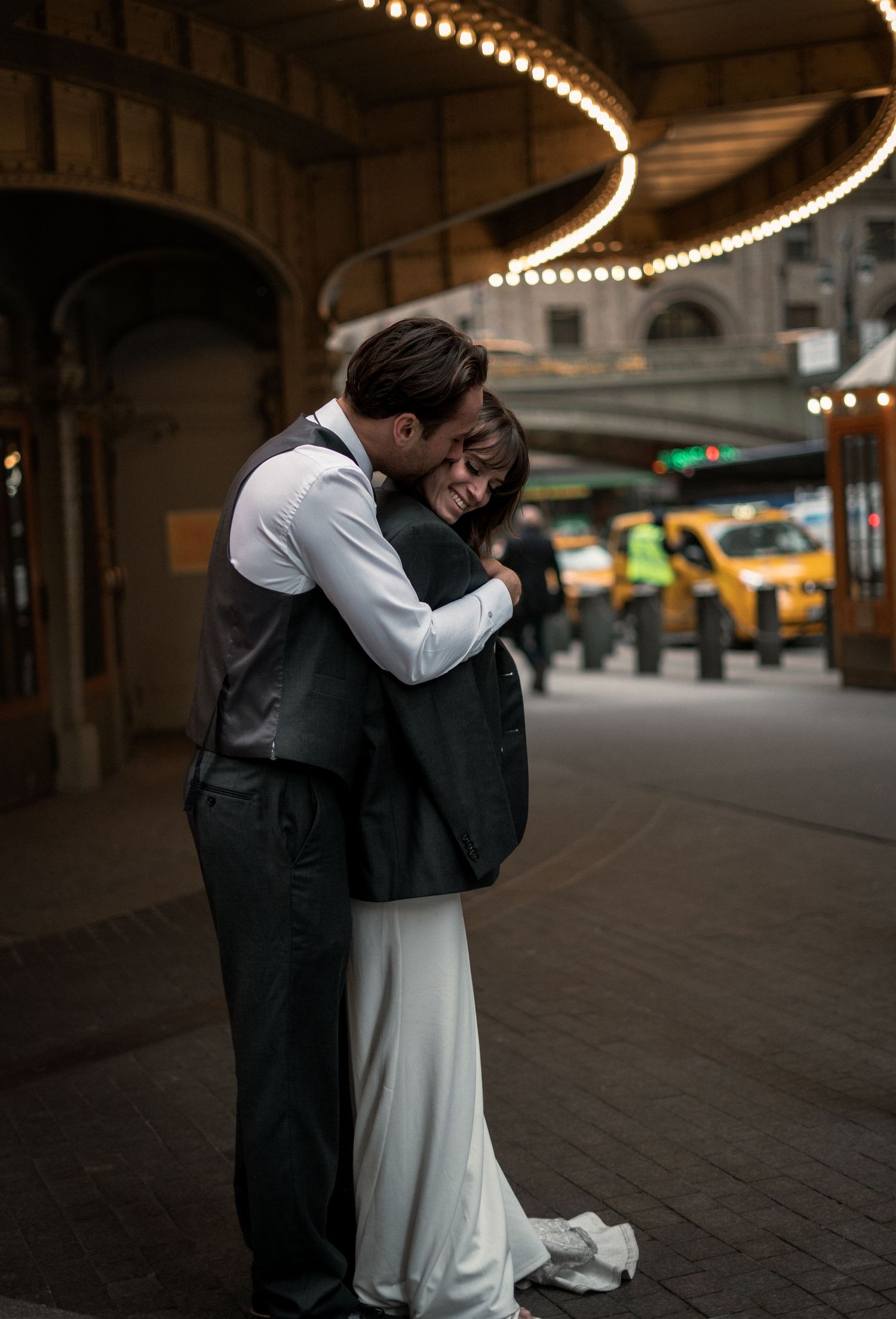 A romantic couple embraces under illuminated archways with yellow taxis visible in the background on a city street.