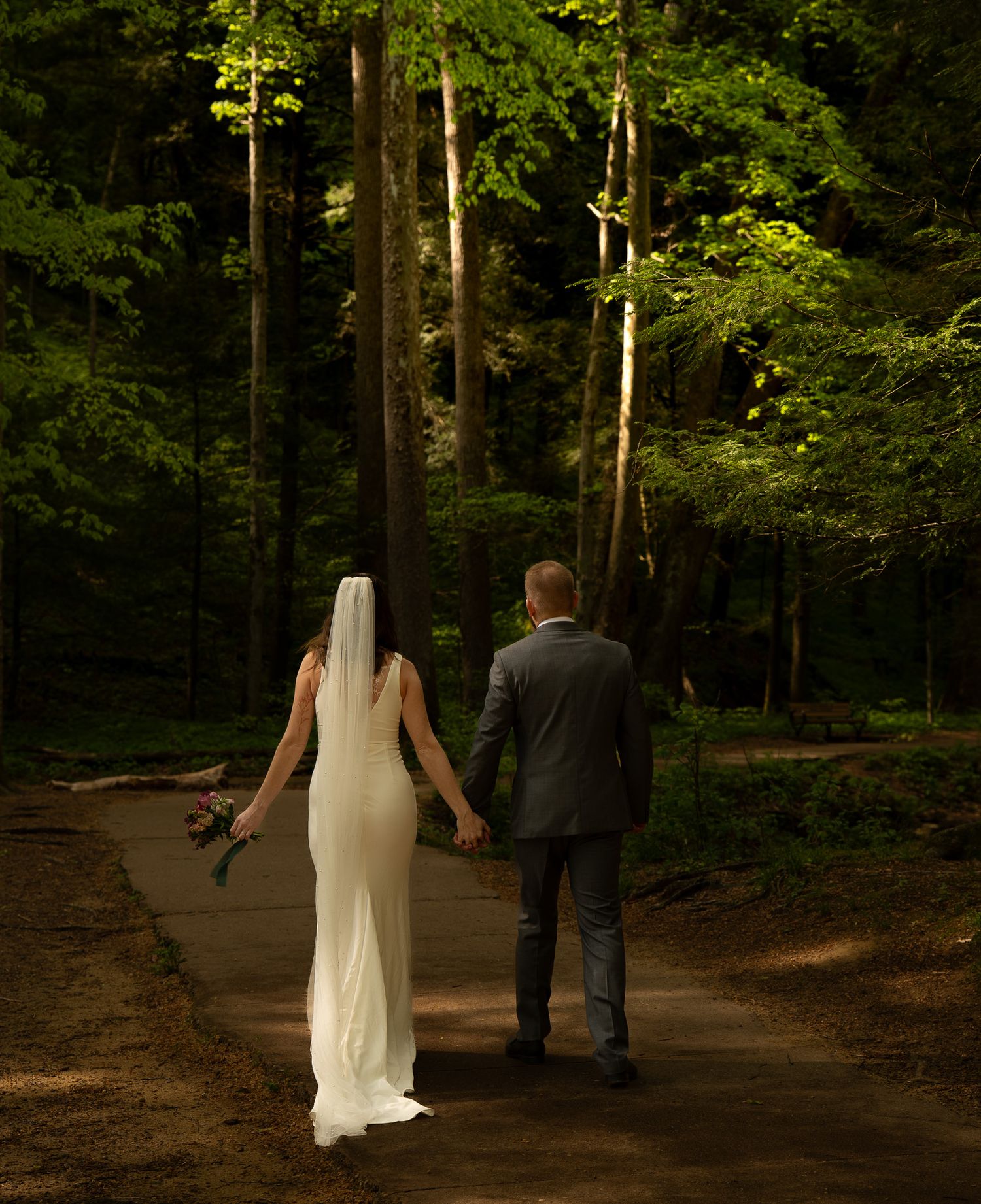 Bride and groom walking hand-in-hand through a sun-dappled forest path, with the bride holding her bouquet behind her.