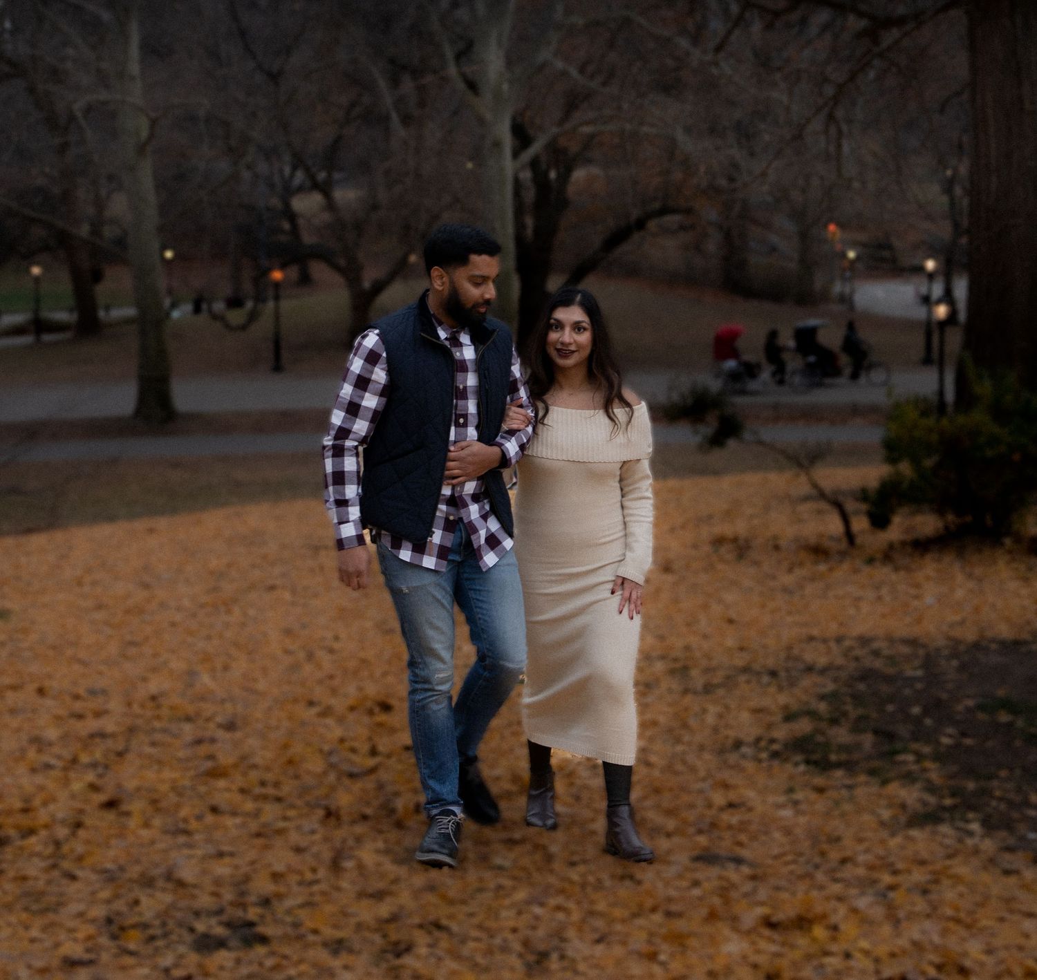 A couple walks together in a park during autumn with fallen leaves on the ground and trees in the background.
