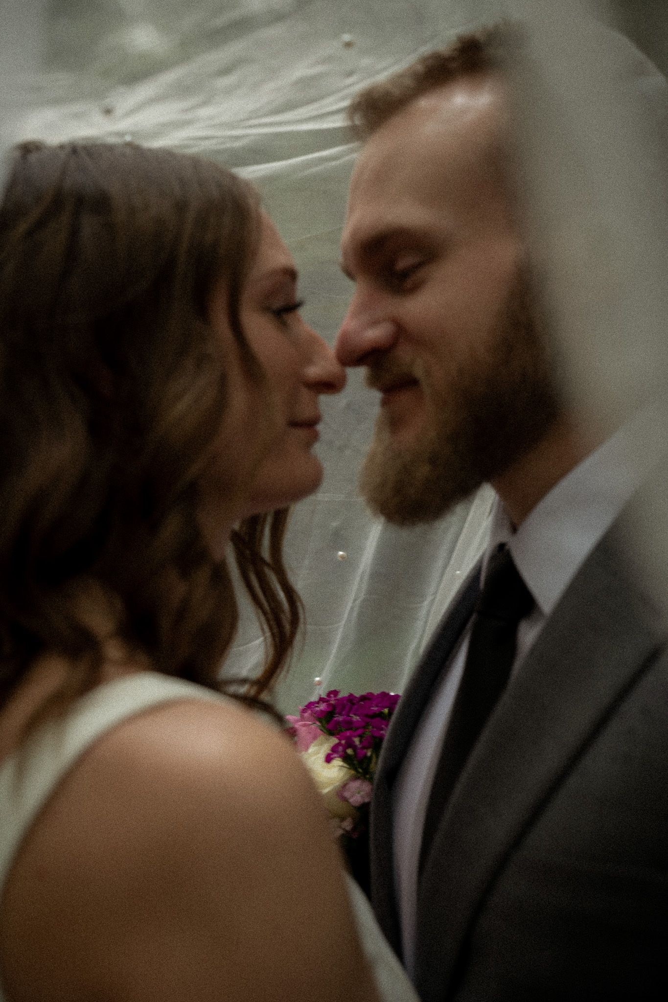 Close-up of bride and groom sharing a quiet moment under the veil, faces gently touching in soft, romantic light.