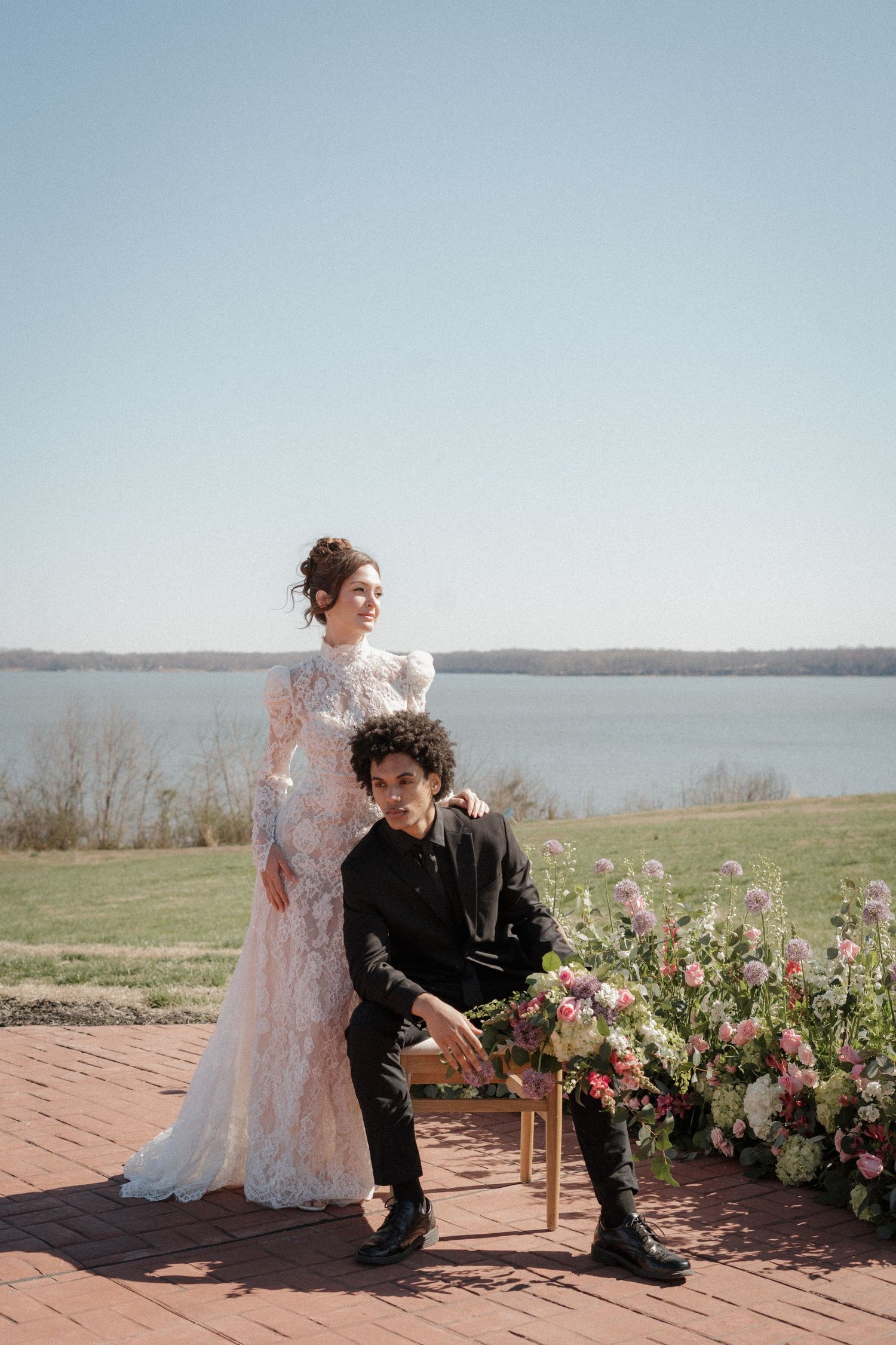 A couple poses on a brick patio overlooking a lake with beautiful floral arrangements at sunset.