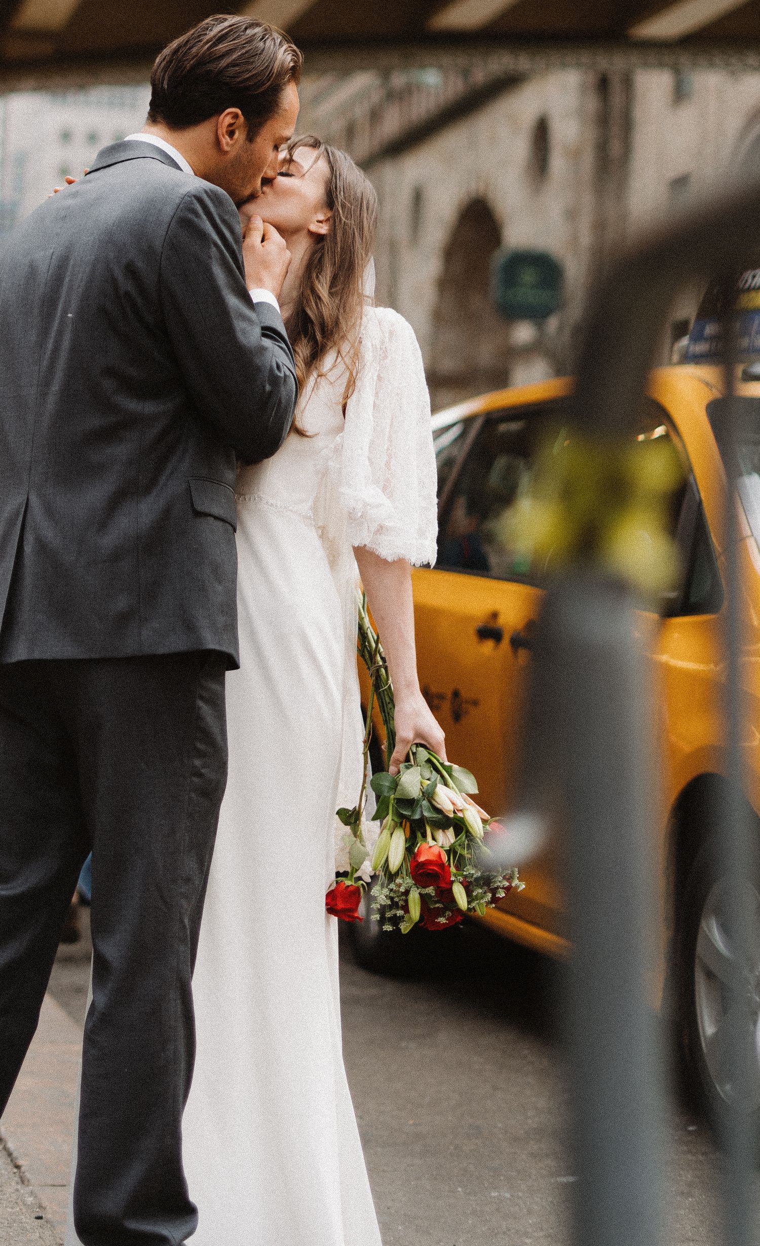 A romantic couple shares a moment on a city street with yellow taxis in the background as the bride holds red roses.