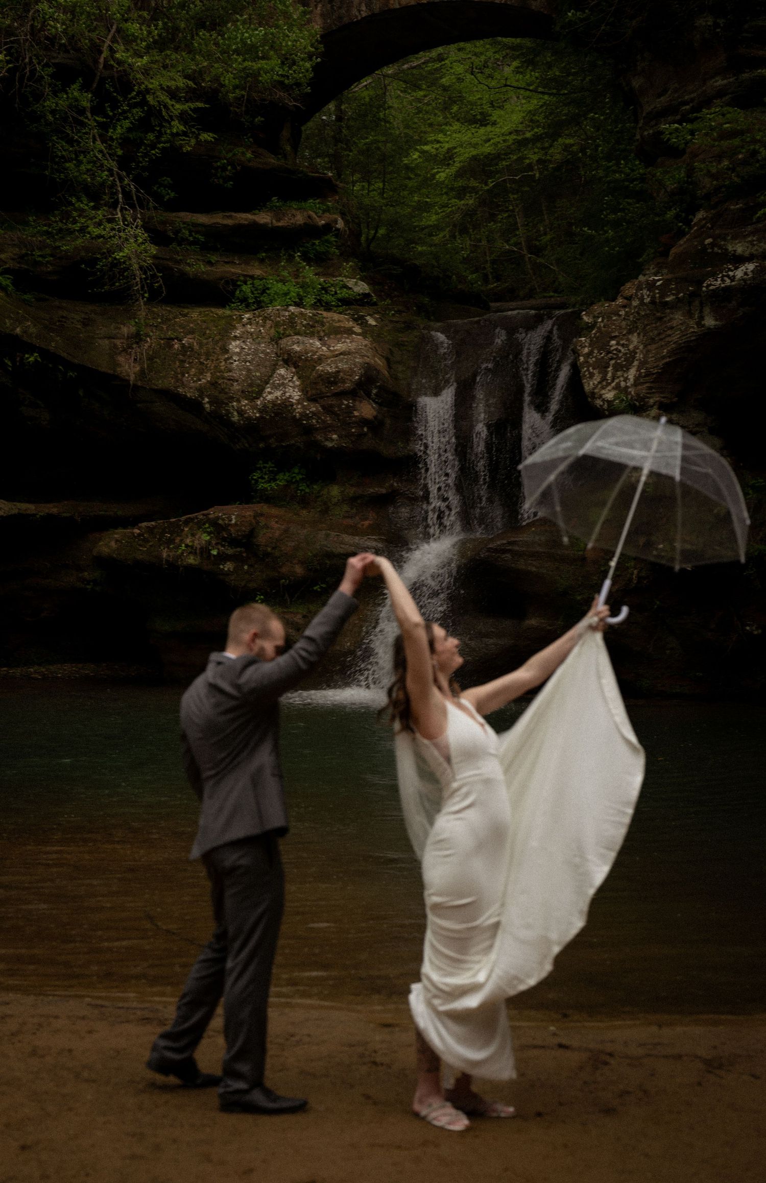 destination national park wedding. Bride and groom dancing playfully near a forest waterfall, the bride lifting her veil with joy.