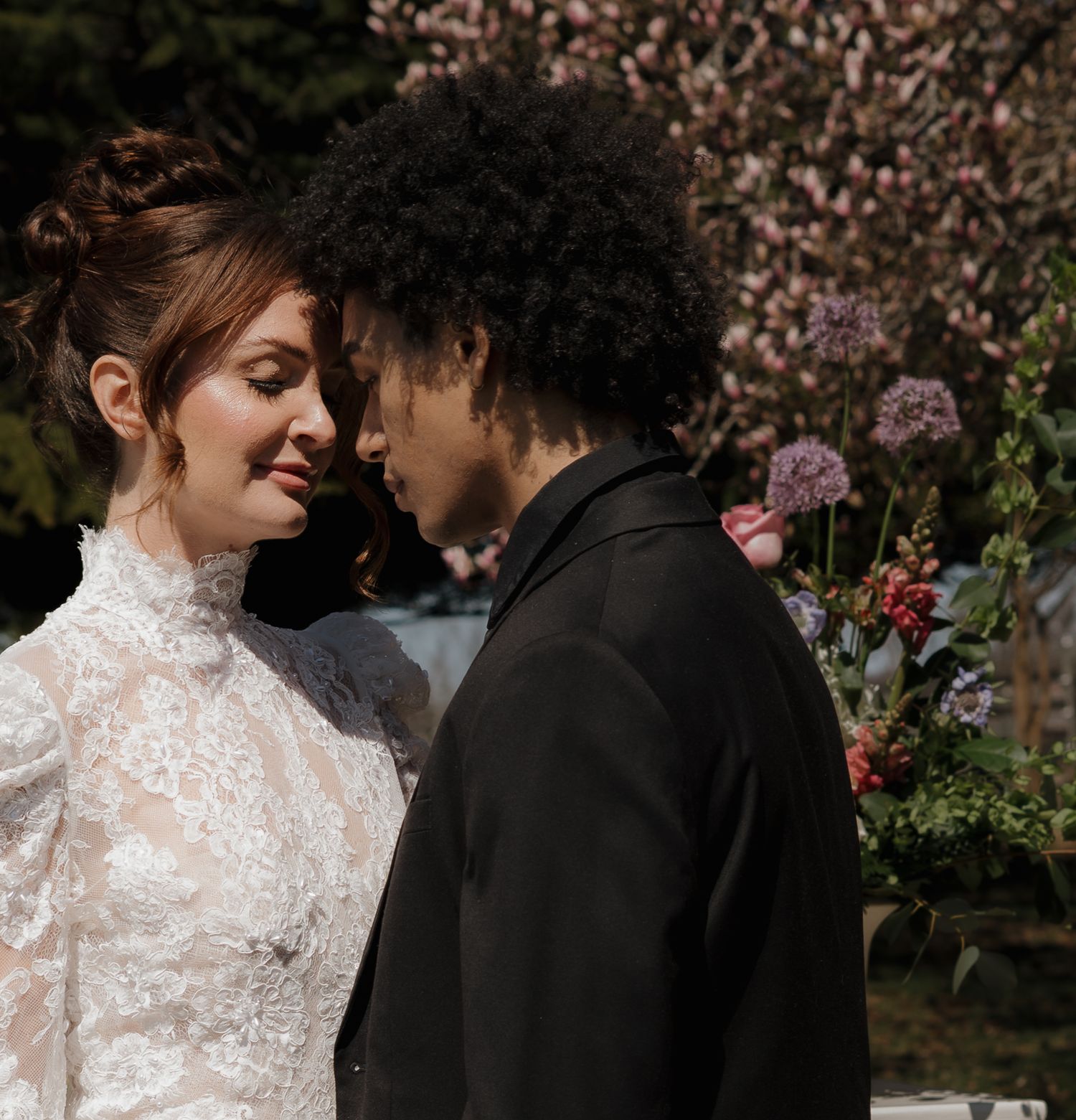 A romantic couple in wedding attire sharing an intimate moment among blooming flowers and garden foliage.